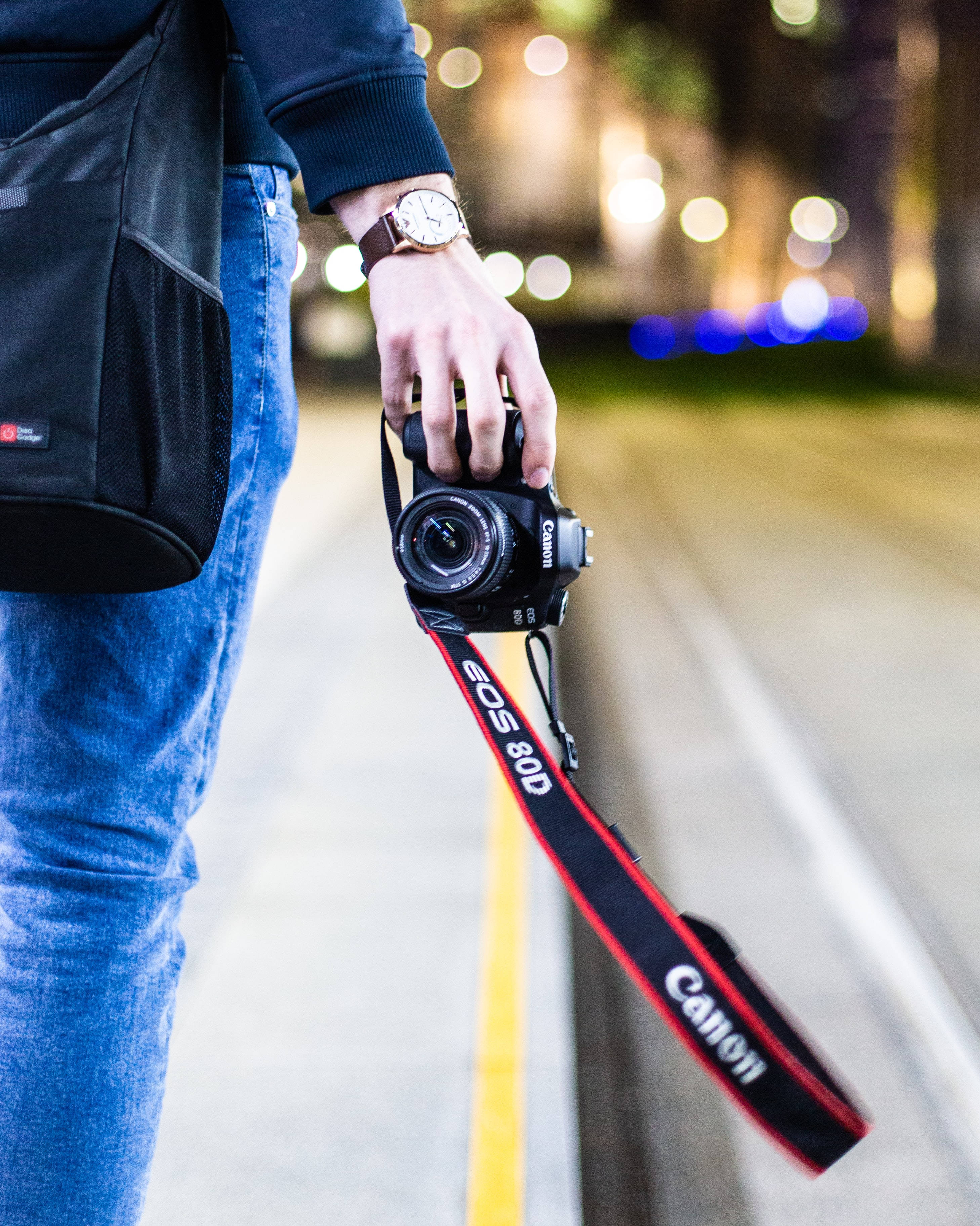 相機 桌布 壁紙 — 一個穿牛仔褲的人夜晚手持Canon EOS 80D相機，背景有散景燈光閃爍。