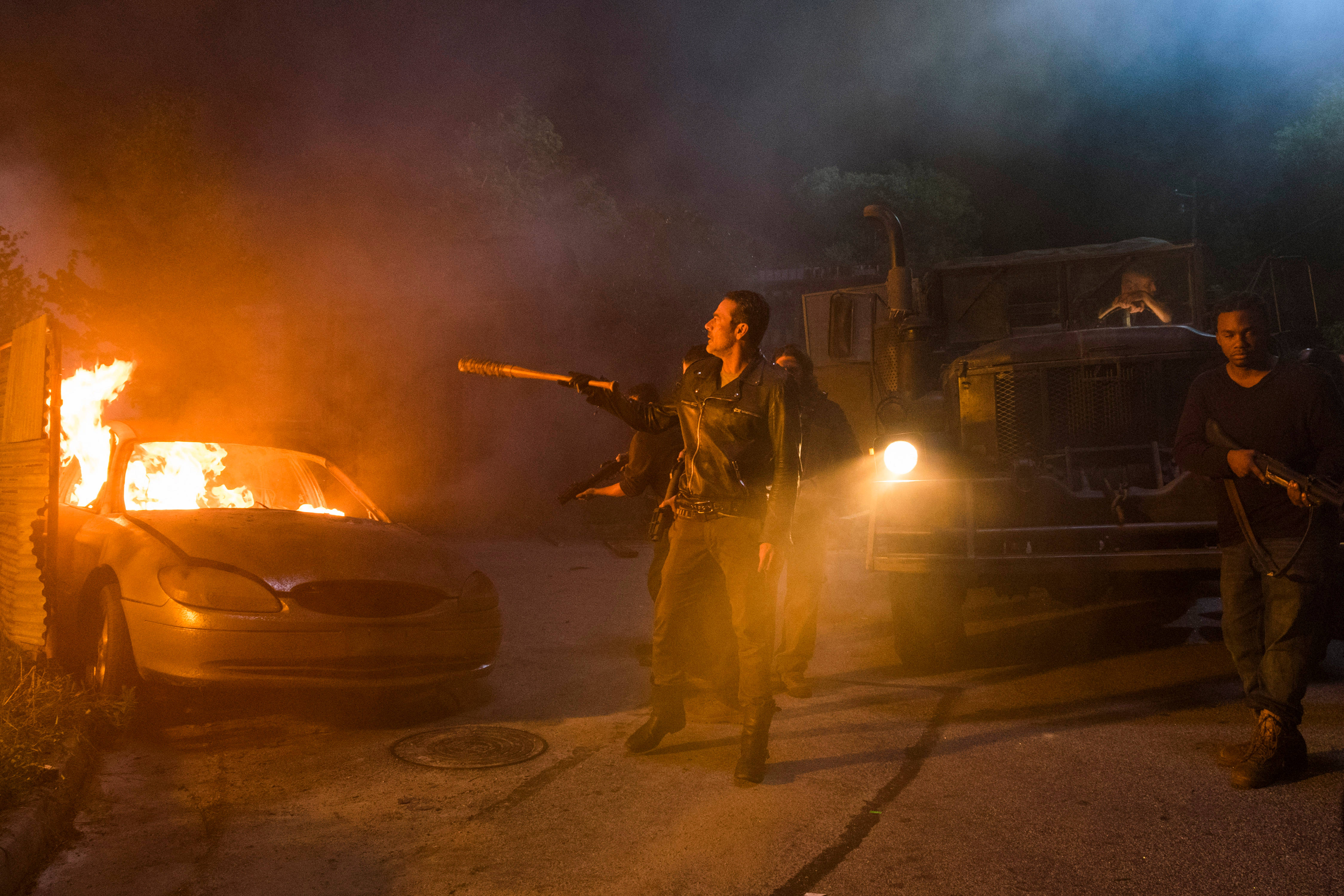 Negan 桌布 壁紙 — 一個穿皮衣的男人用步槍瞄準一輛燃燒的汽車，煙霧在夜晚卡車頭燈的光芒下翻騰。
