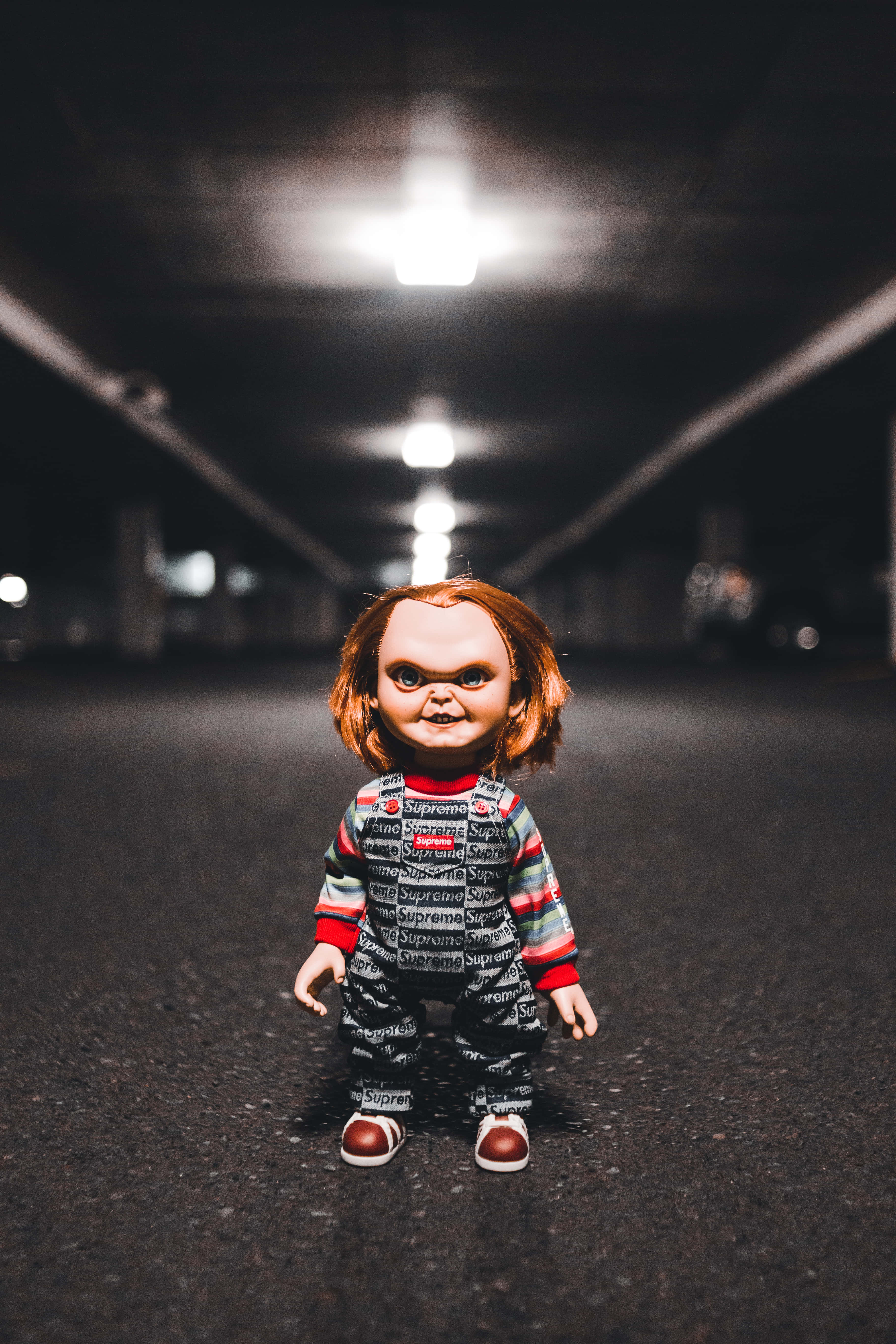 鬼娃恰吉 桌布 壁紙 — 一個穿著 Supreme 服裝的 Chucky 娃娃獨自站在光線昏暗的隧道中，頭頂有發光的燈光。