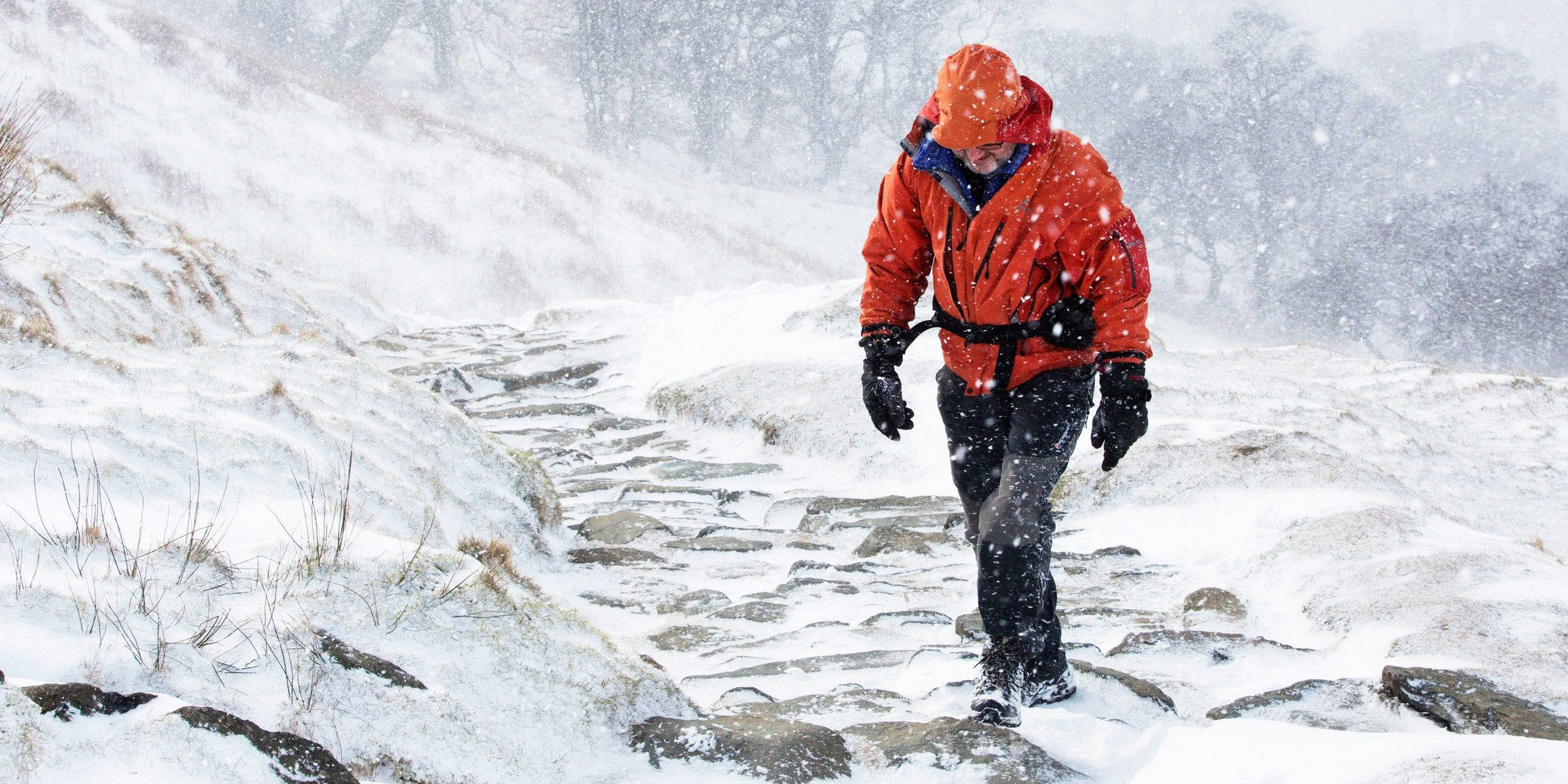 冬季 桌布 壁紙 — 一個穿著亮橙色外套的人正在雪覆蓋的山徑上徒步，雪花在他們周圍飄落。