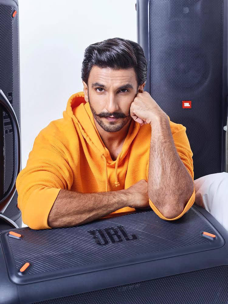Ranveer Singh 桌布 壁紙 — 一個穿著亮橙色連帽衫的男人若有所思地靠在他身後帶有大揚聲器的黑色JBL揚聲器上。
