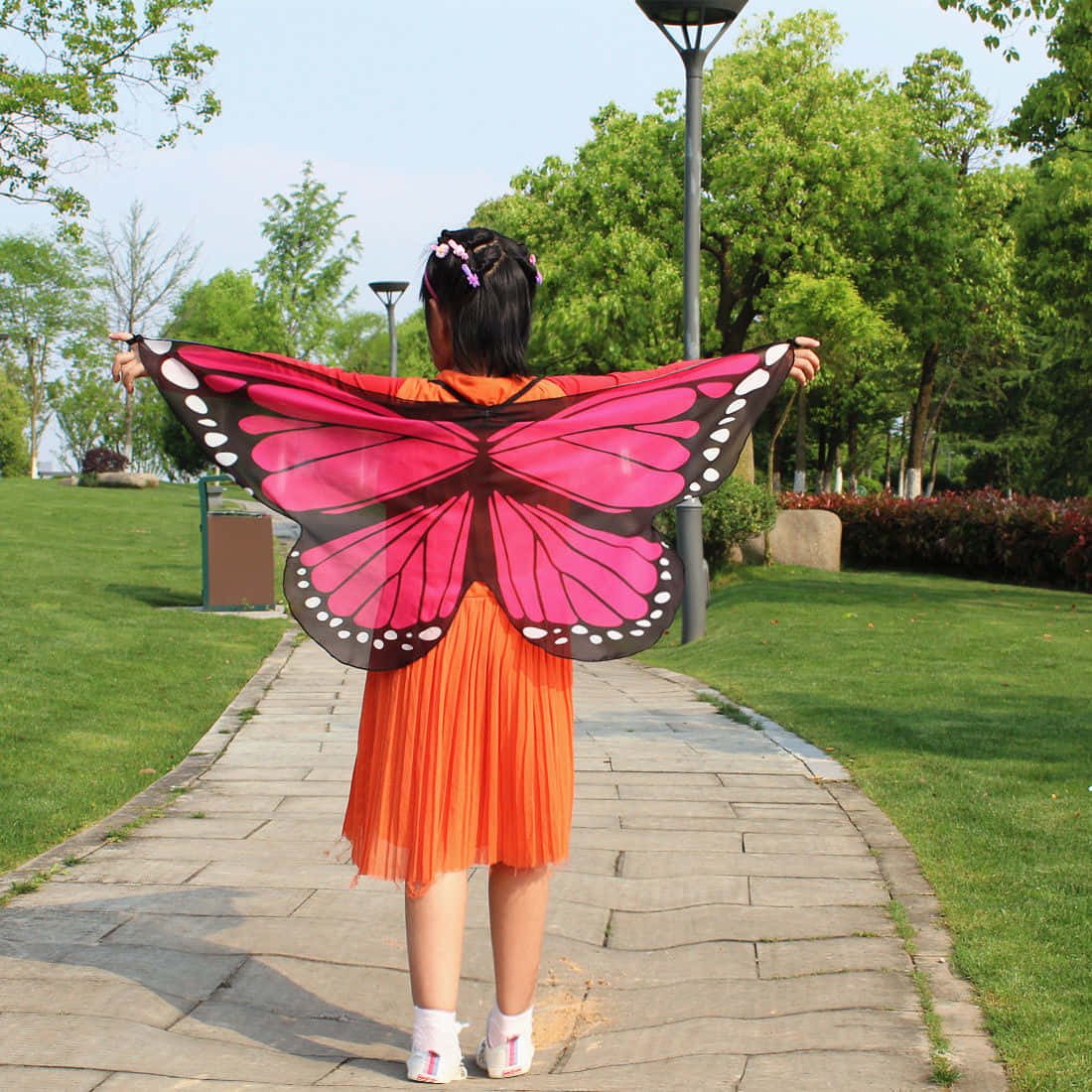 Butterfly Wing Dress 桌布 壁紙 — 一個穿著亮橙色連衣裙的孩子，戴著粉紅色和黑色的大蝴蝶翅膀，沿著公園小徑走遠。