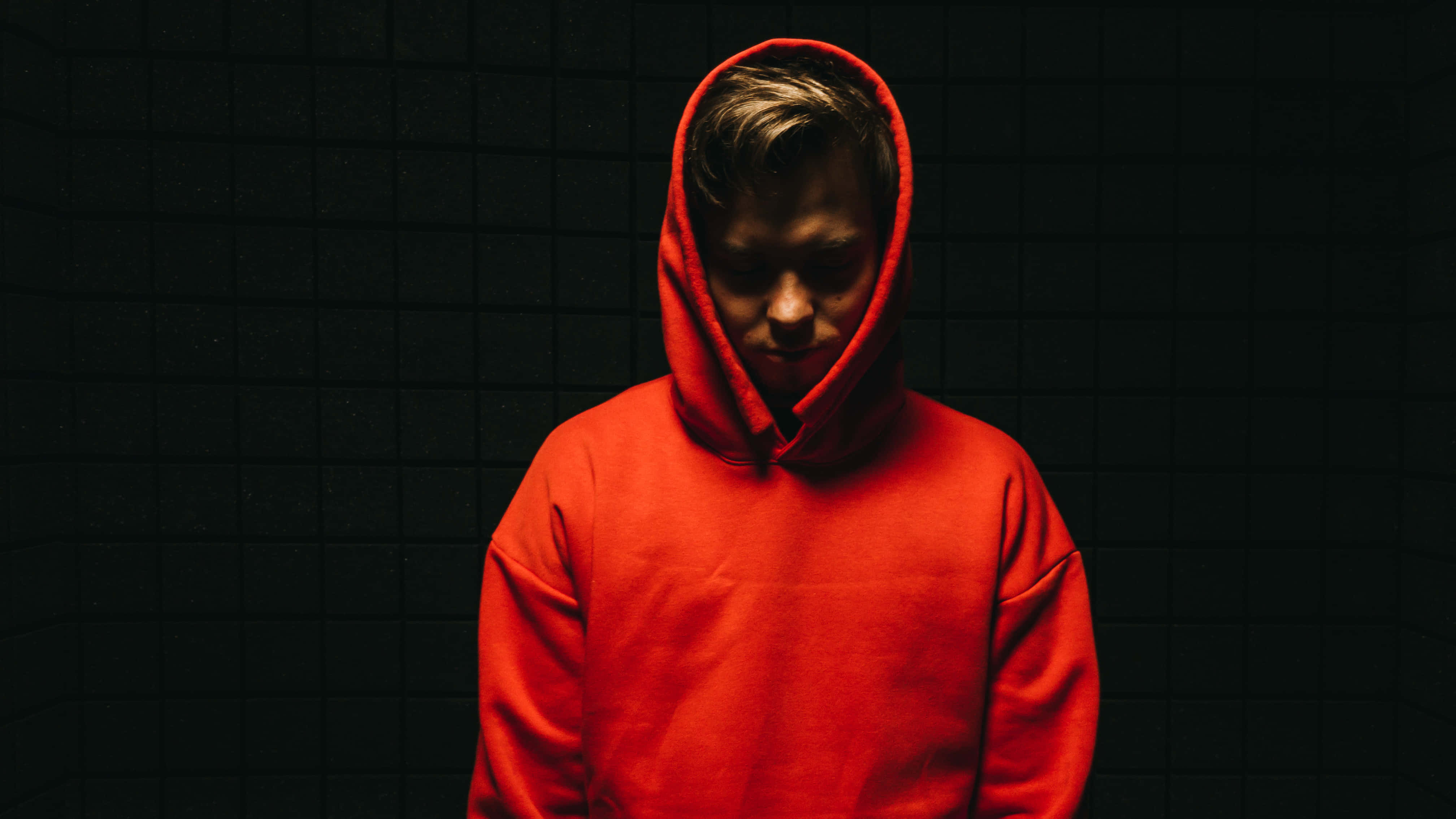 Red Hoodie 桌布 壁紙 — 一個穿著亮紅色連帽衫的人站在深色瓷磚牆前，低調的燈光在他們的臉上投下陰影。