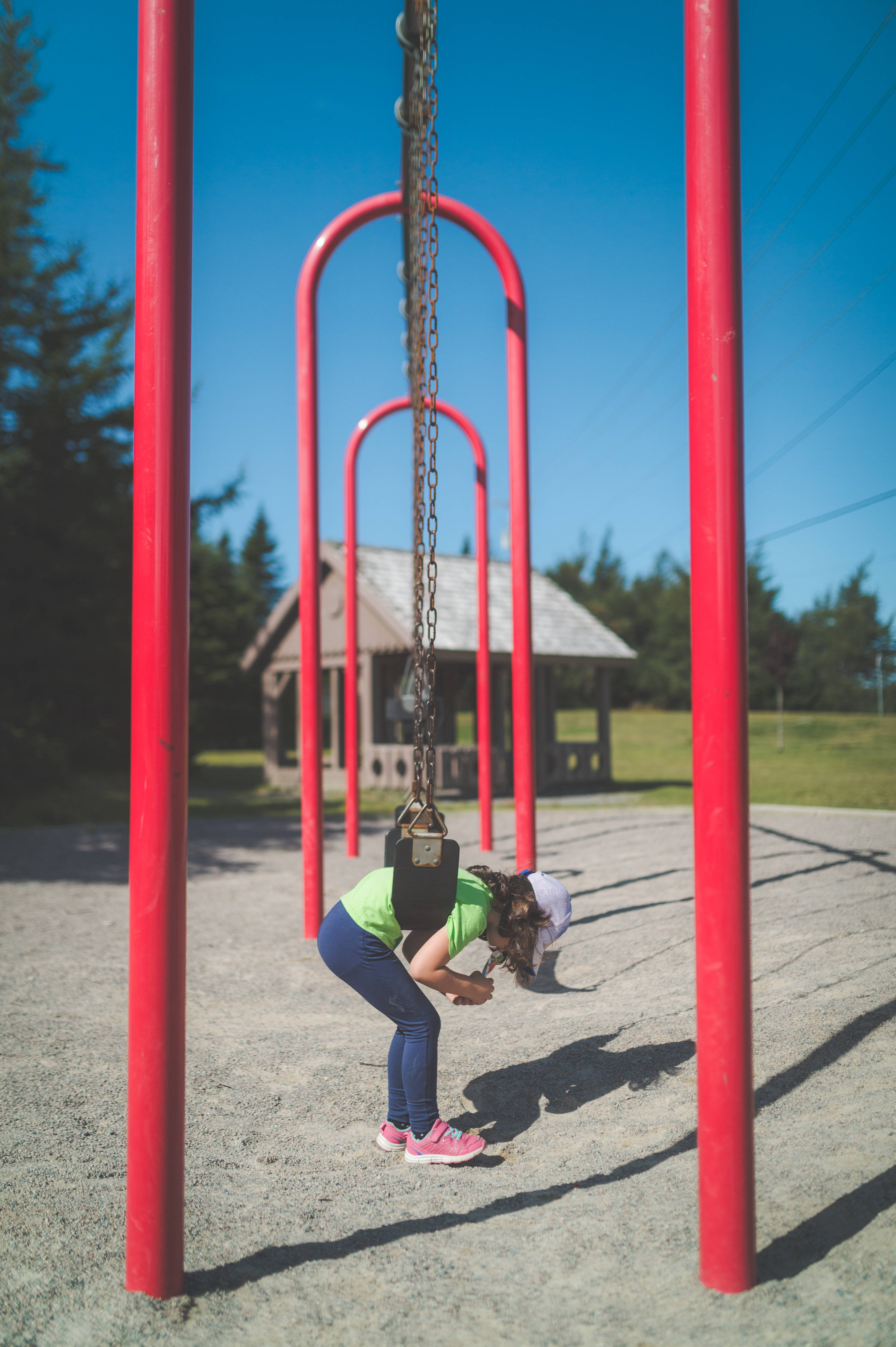 Playground 桌布 壁紙 — 一個穿著亮綠色襯衫的小孩在陽光明媚的遊樂場上，彎腰在紅色鞦韆架下繫鞋帶，背景有一個木製遮陽棚。