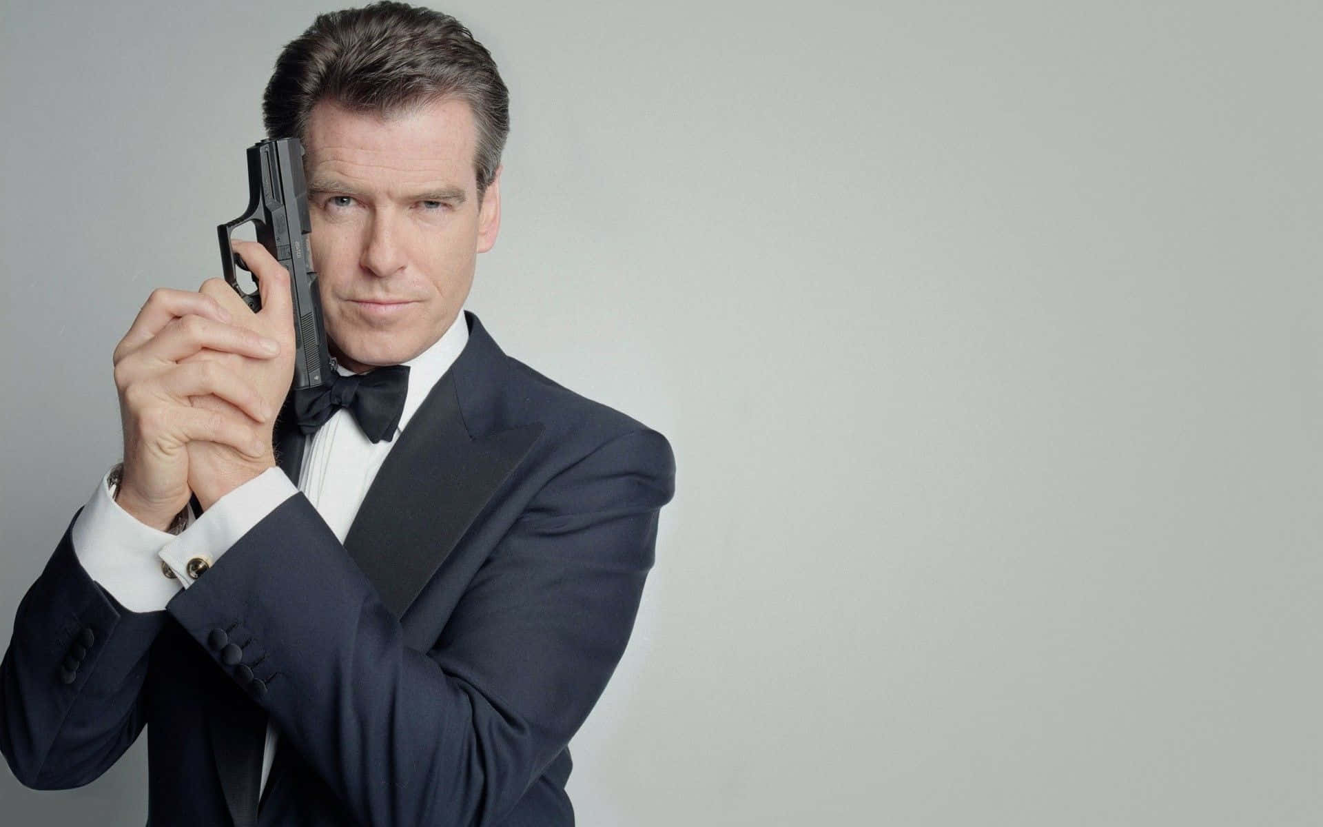pierce brosnan 桌布 壁紙 — 一個穿著俐落黑色燕尾服的男人，在純灰色背景下，手持手槍，神情嚴肅。