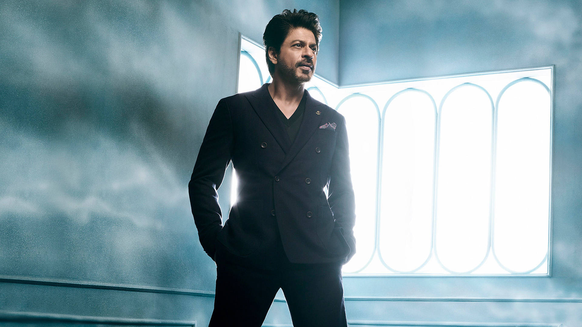 Shah Rukh Khan 桌布 壁紙 — 一個穿著俐落黑色西裝的男人，雙手插袋，自信地站在一個帶有明亮拱形窗戶的憂鬱藍色雲彩背景前。