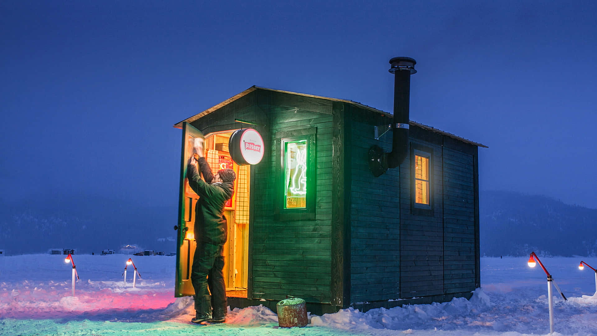 Ice Fishing 桌布 壁紙 — 一個穿著冬季裝備的人，在雪景暮色中打開一間溫暖照明的綠色木屋門。