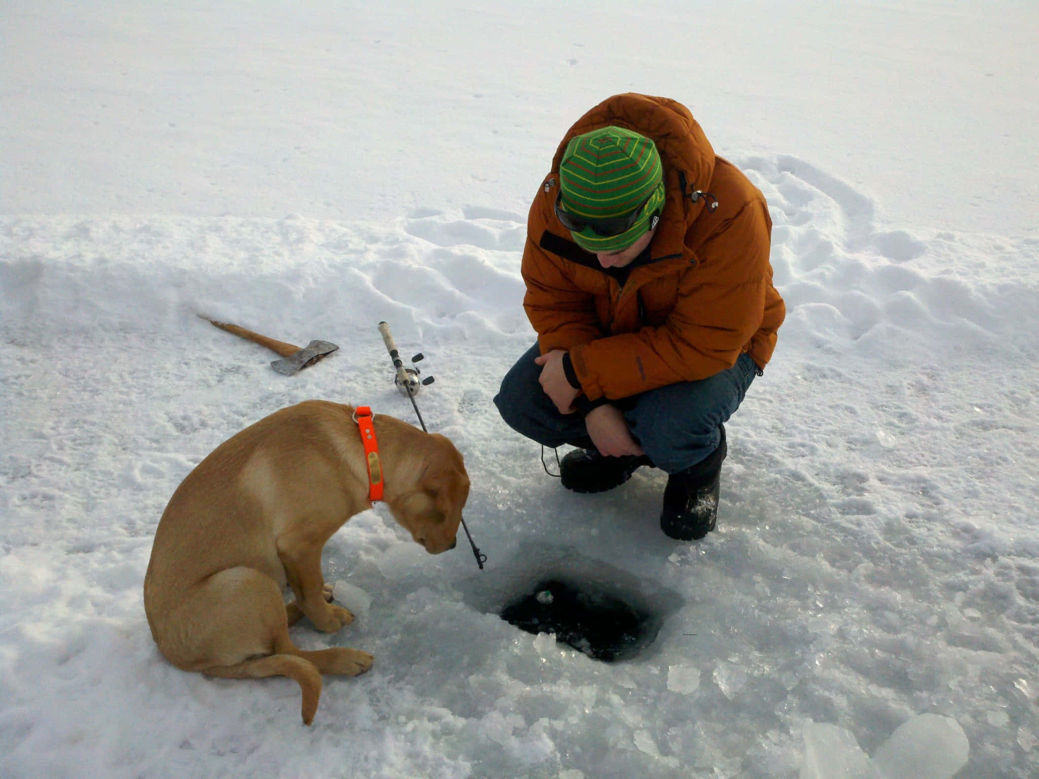 Ice Fishing 桌布 壁紙 — 一個穿著冬季裝備的人在雪景中蹲在狗旁邊，靠近一個釣魚洞。