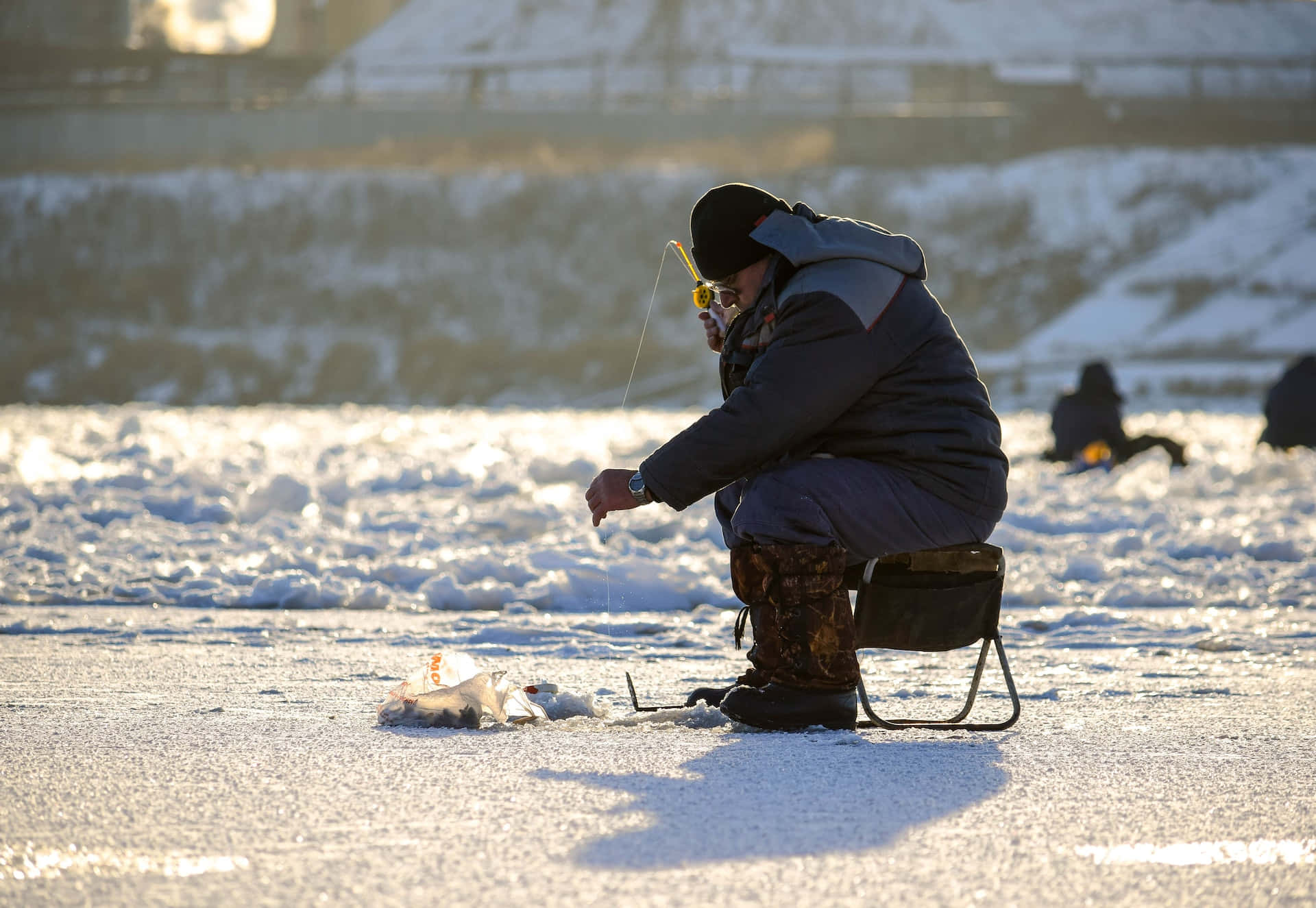 Ice Fishing 桌布 壁紙 — 一個穿著冬季裝備的人坐在冰凍湖面上的凳子上，在黃金時段將釣線穿過冰洞放下。