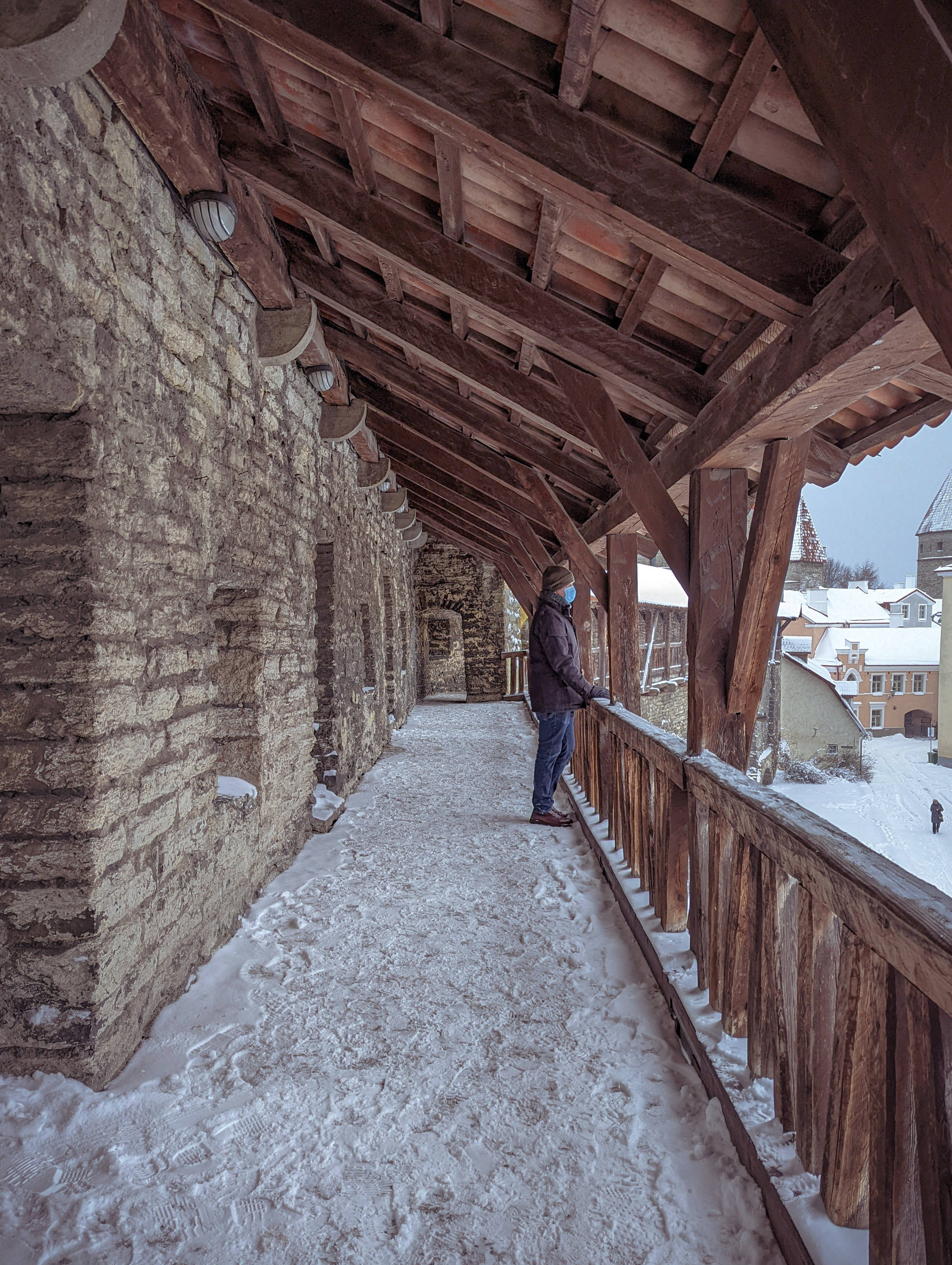 Estonia 桌布 壁紙 — 一個穿著冬季裝備的人站在一條歷史悠久的石橋上，雪覆蓋的木製步道上，俯瞰一個被雪覆蓋的小鎮。