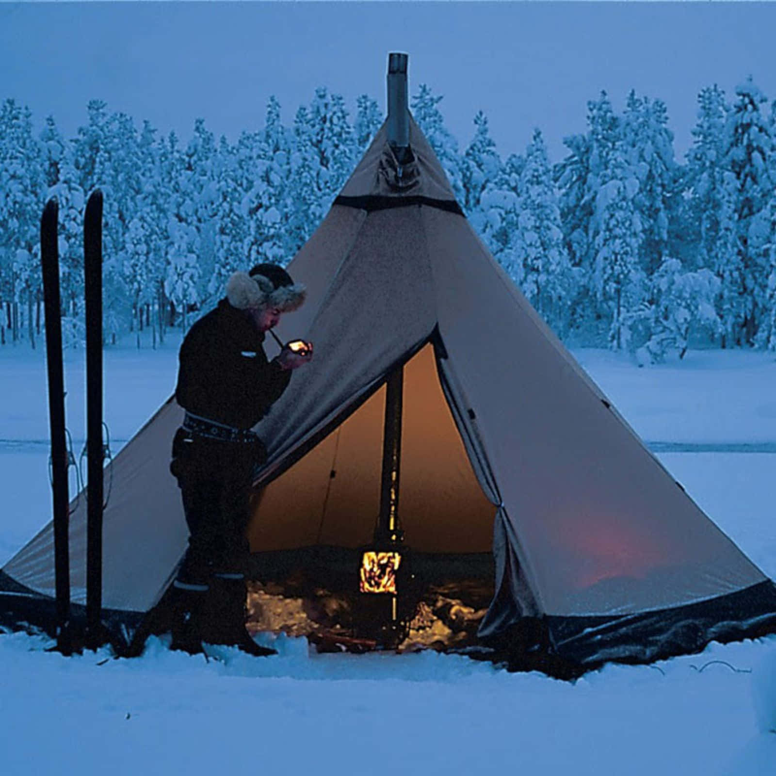 Winter Camping 桌布 壁紙 — 一個穿著冬季裝備的人站在黃昏時分被雪覆蓋的帳篷旁，手持燈籠，溫暖的光線從內部流出，映襯著雪白的森林背景。