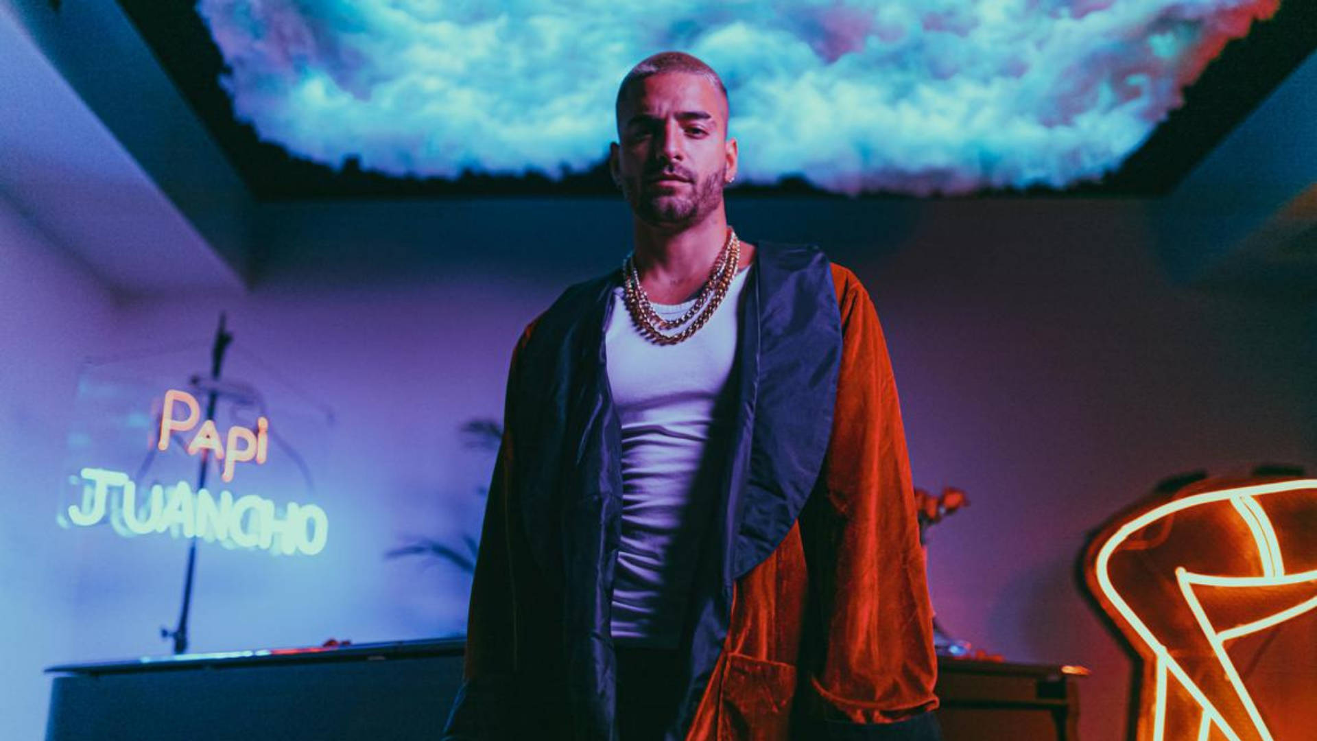 Maluma 桌布 壁紙 — 一個穿著多層次服裝的男人站在發光的雲層天花板下，霓虹燈牌寫著 Papi Juancho。