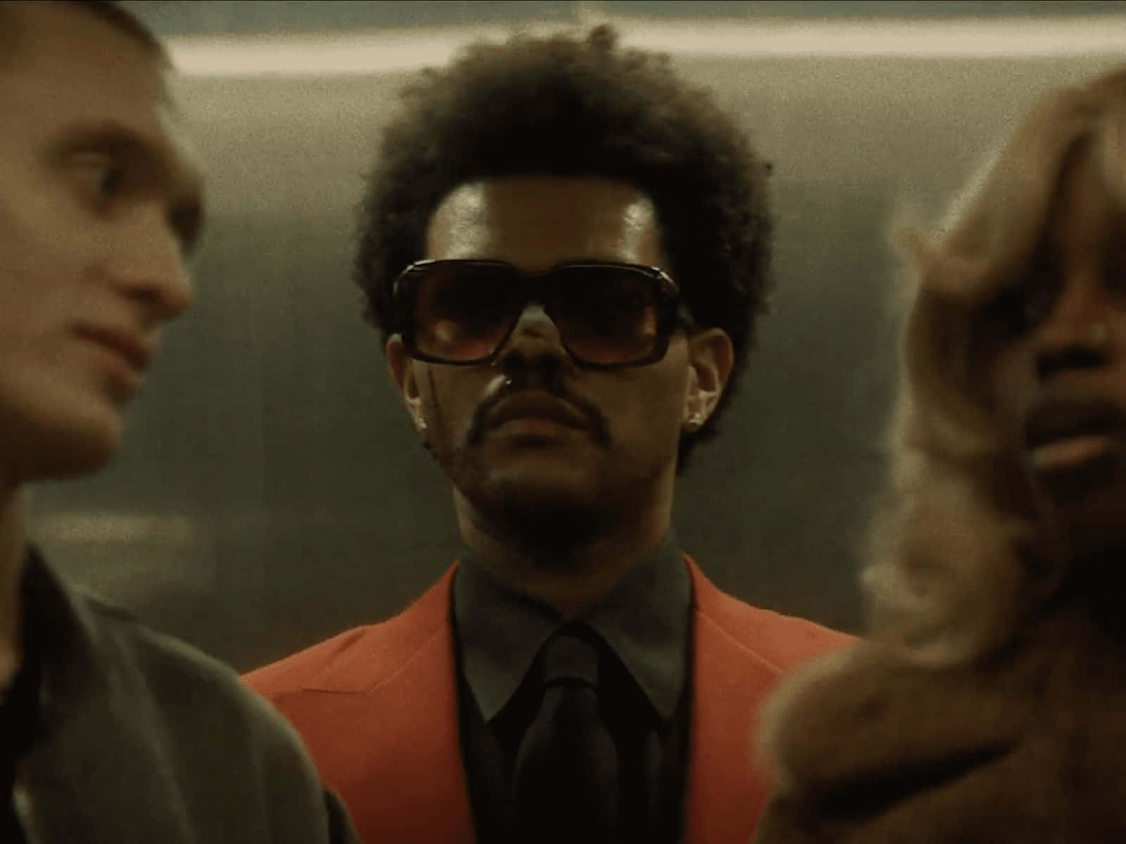 The Weeknd After Hours 桌布 壁紙 — 一個穿著大膽橙色西裝和超大太陽眼鏡的男人站在中央，兩側有兩個模糊的人影，在昏暗、充滿氛圍的室內。
