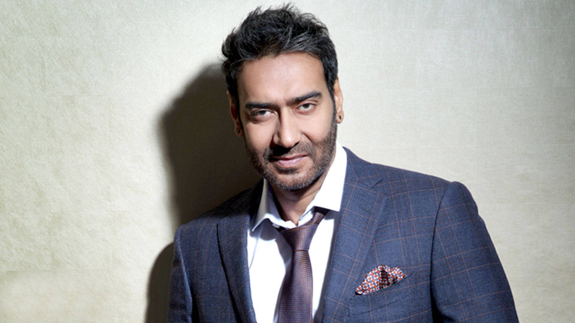 ajay devgn 桌布 壁紙 — 一個穿著尖銳格紋西裝的男人靠在一面有紋理的牆上，戲劇性的燈光投射出陰影。