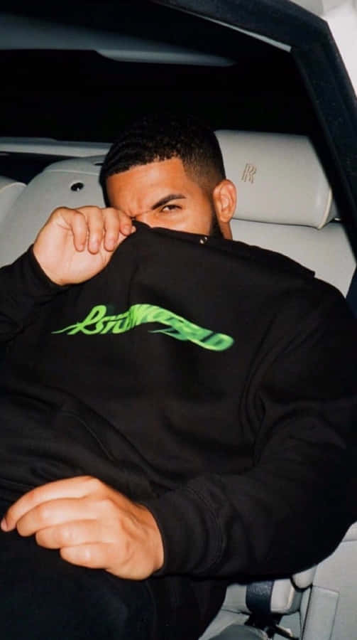 Aesthetic Drake 桌布 壁紙 — 一個穿著帶有綠色文字的黑色連帽衫的男人坐在豪華車裡，拉起帽子遮住臉。