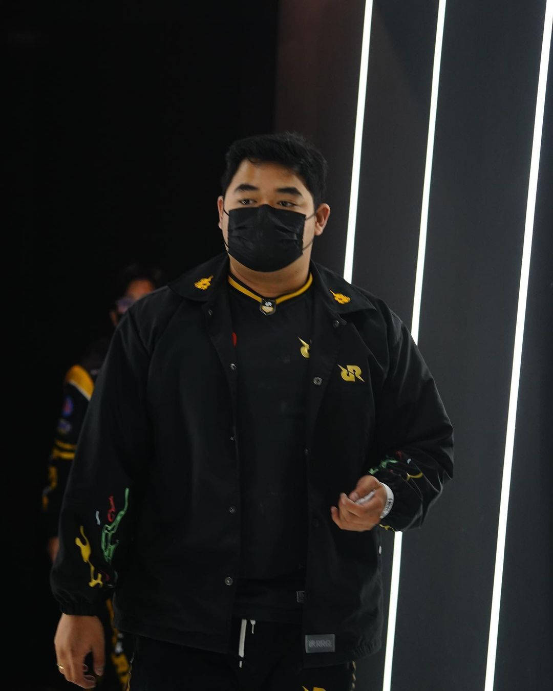Team 桌布 壁紙 — 一個穿著帶有黃色和綠色點綴的黑色外套的人，站在有明亮垂直光帶的深色牆壁前。
