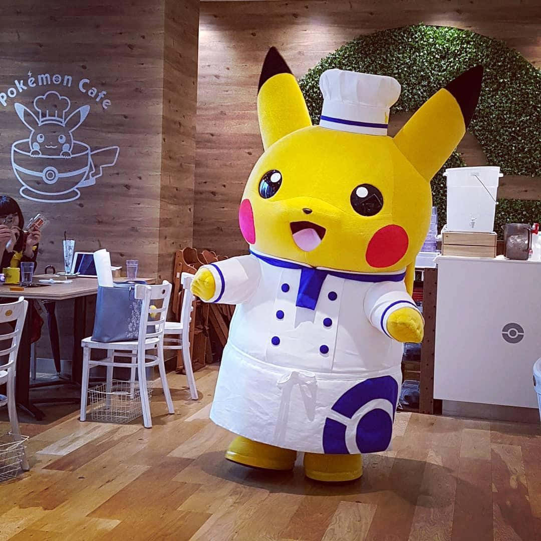 Tokyo Japan 桌布 壁紙 — 一個穿著廚師服裝的開心皮卡丘吉祥物站在一個有木牆和附近有顧客用餐的寶可夢主題咖啡館裡。