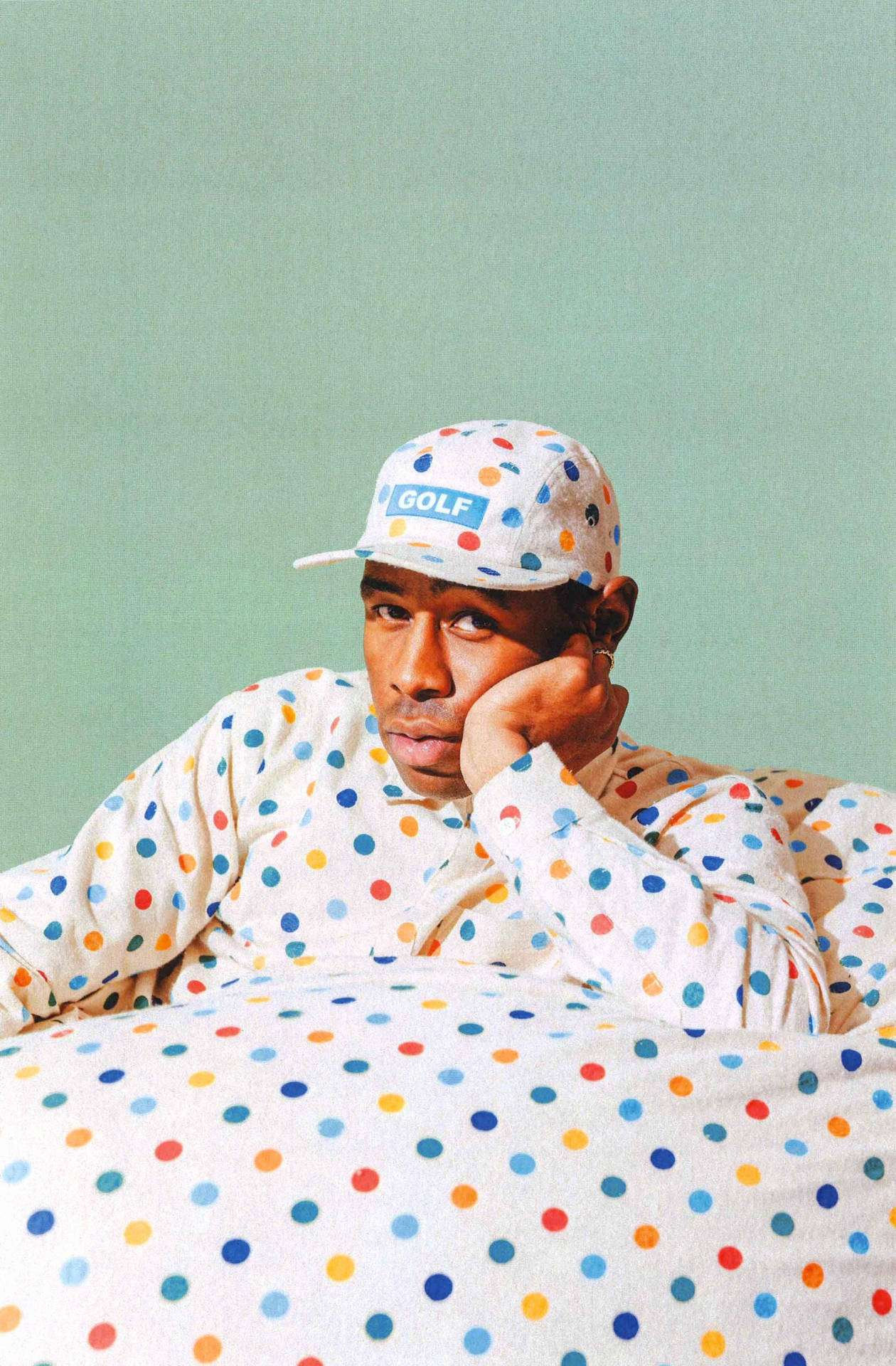 tyler 桌布 壁紙 — 一個穿著彩色圓點服裝和帽子的男人，在柔和的綠色背景前將頭靠在手上。