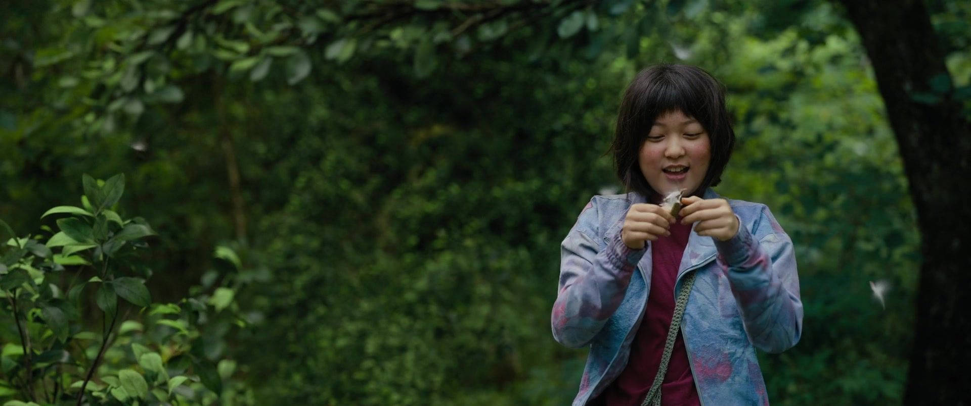 okja 桌布 壁紙 — 一個穿著彩色外套的年輕女孩在茂密的綠色森林中微笑，手裡拿著一個小物件。