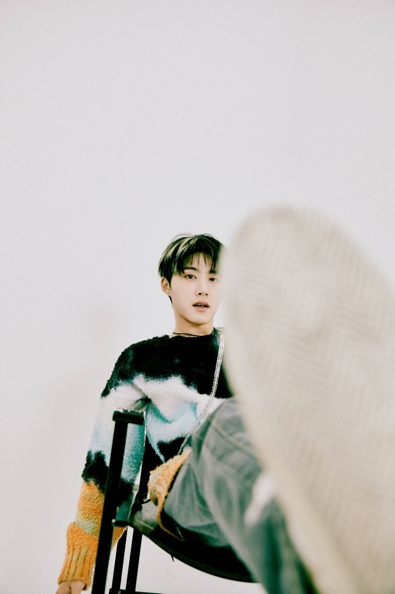 kim hanbin 桌布 壁紙 — 一個穿著彩色毛絨毛衣的年輕男子坐在椅子上，背景是純白色，前景有一個模糊的紋理物體。
