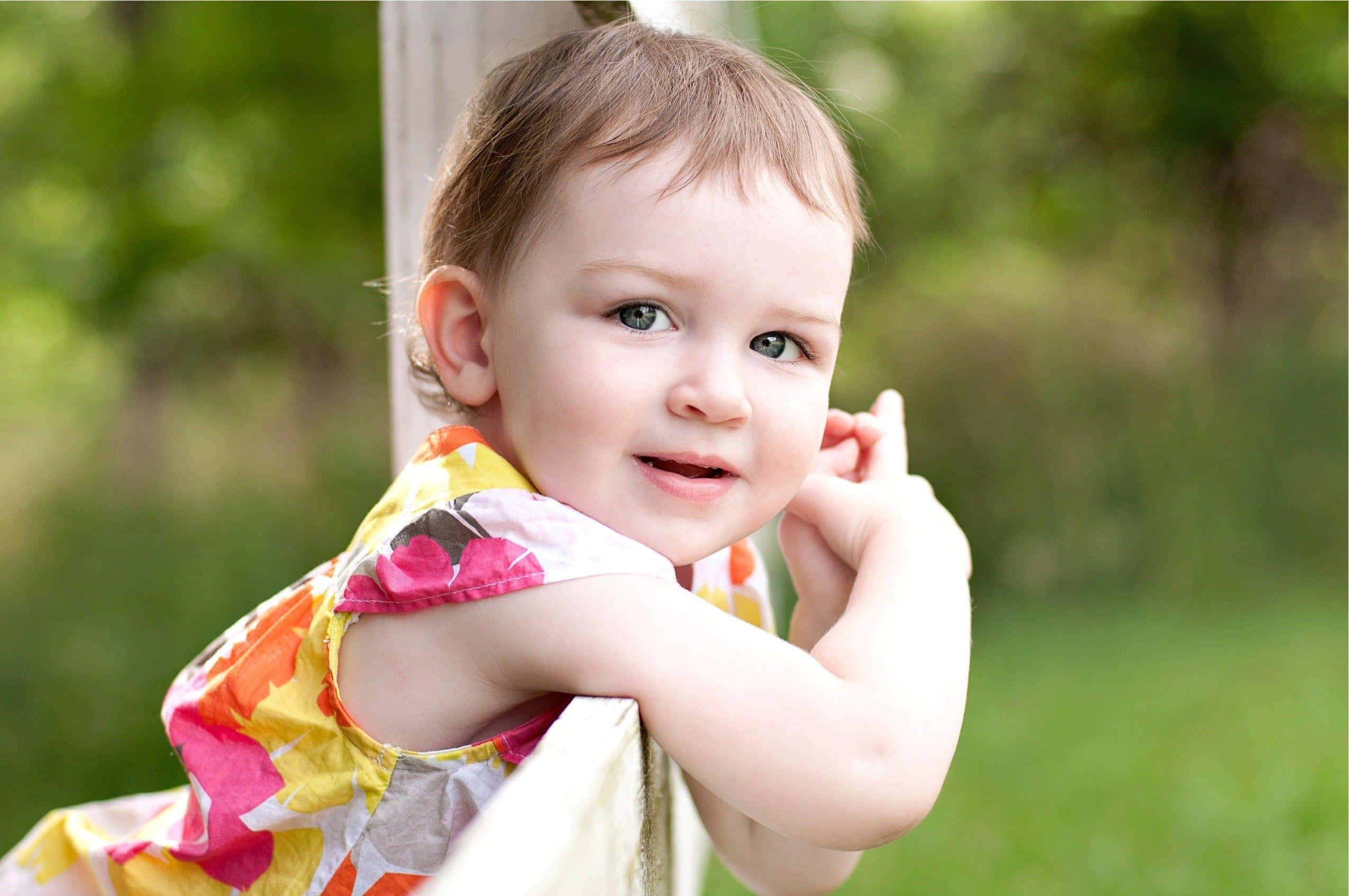 Baby Girl 桌布 壁紙 — 一個穿著彩色花卉連衣裙的開朗幼兒靠在木柵欄上，背景是明亮的綠色模糊效果。