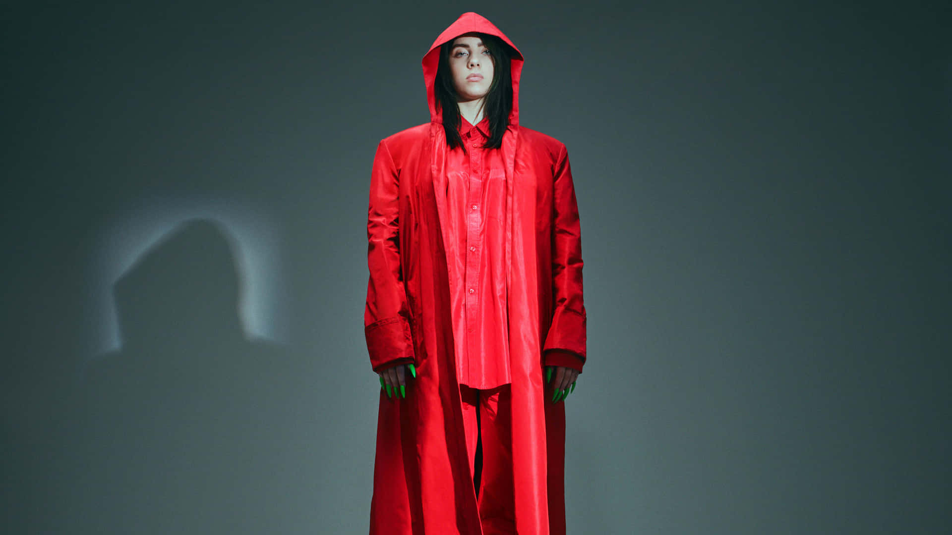 Billie Eilish Laptop 桌布 壁紙 — 一個穿著搶眼紅色連帽裝的人站在灰色牆壁前，身後投射出戲劇性的陰影。
