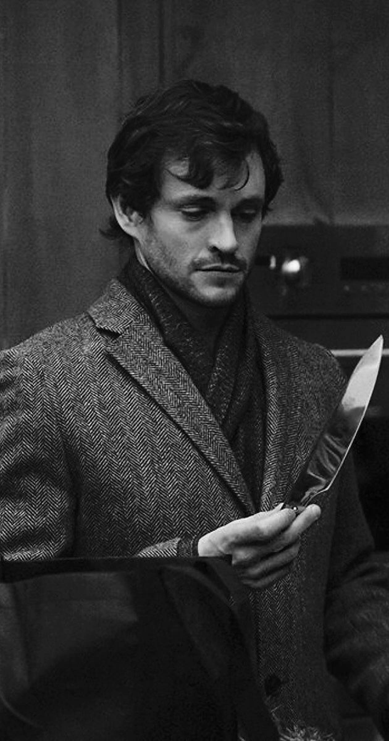 Hugh Dancy 桌布 壁紙 — 一個穿著有紋理外套的男人在昏暗的房間裡專注地拿著一把刀。