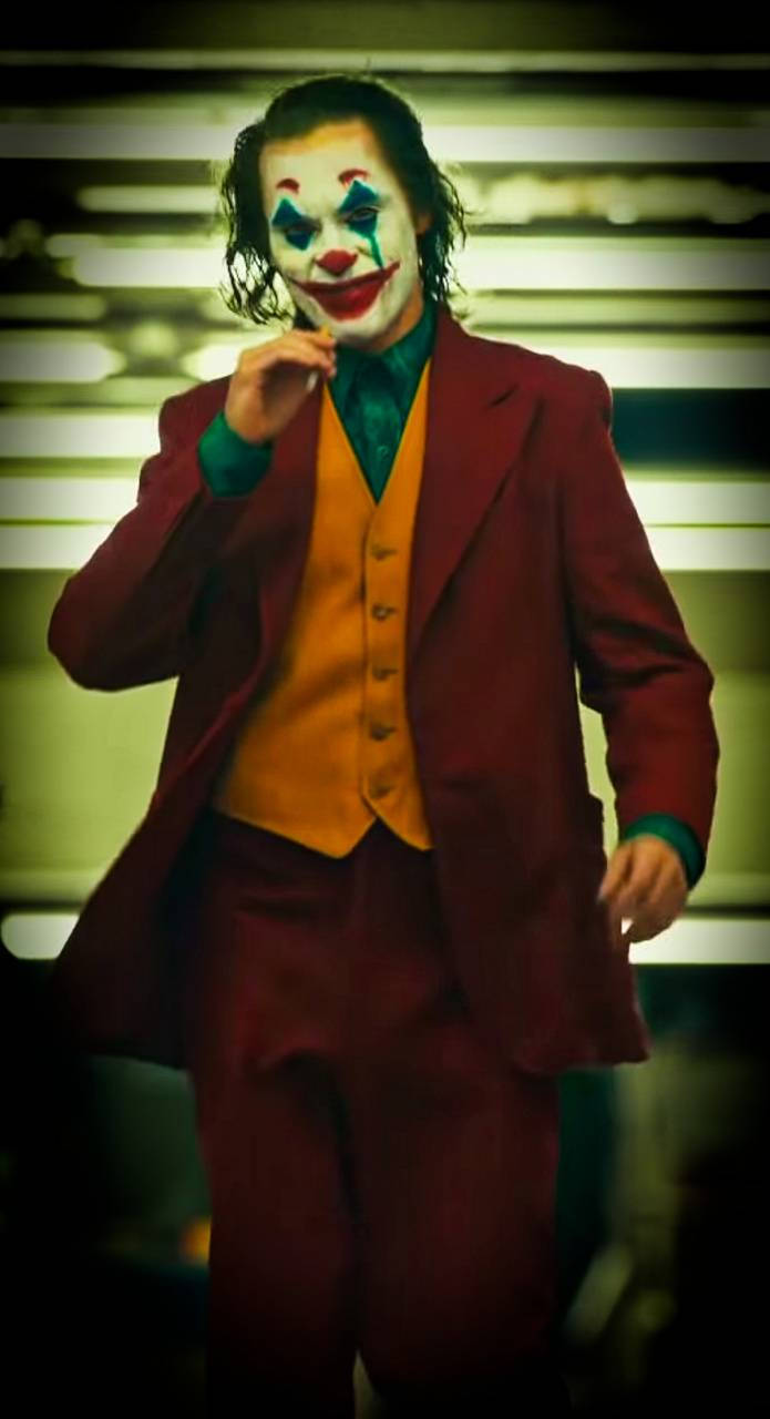 Joker 2019 桌布 壁紙 — 一個穿著栗色西裝和橙色背心的可怕小丑，站在模糊的條紋背景前，燈光效果戲劇性。