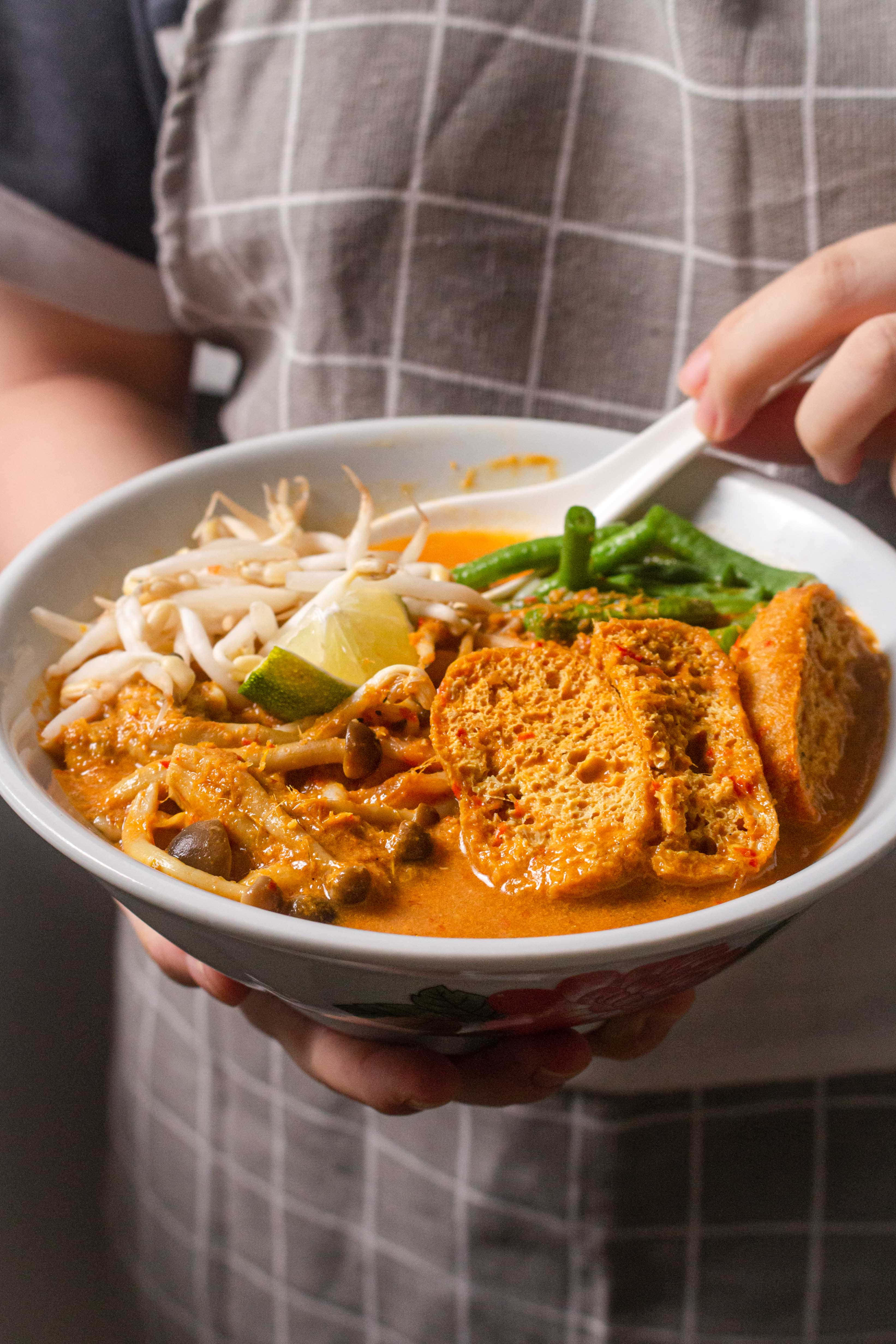 vegan 桌布 壁紙 — 一個穿著格子圍裙的人拿著一碗冒煙的濃郁橙色咖哩湯麵，上面有豆腐、豆芽和萊姆。