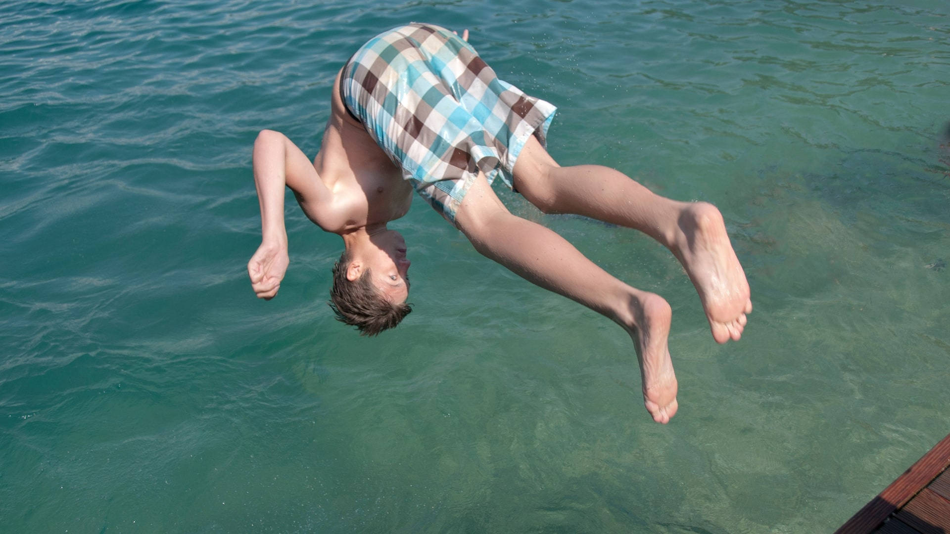 tumbling 桌布 壁紙 — 一個穿著格子短褲的男孩從木碼頭一頭扎進清澈的藍綠色水中。