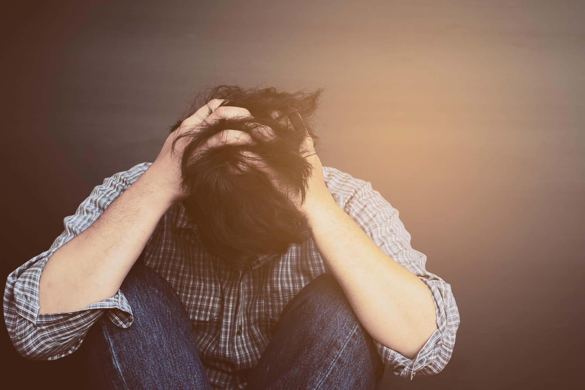 Sad Face 桌布 壁紙 — 一個穿著格子衫的人彎腰坐著，雙手抱頭，背景是漸變棕色。