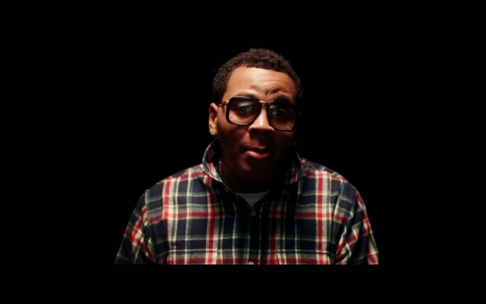 Kevin Gates 桌布 壁紙 — 一個穿著格子襯衫和厚眼鏡的男人，在黑色背景和戲劇性燈光下凝視著。
