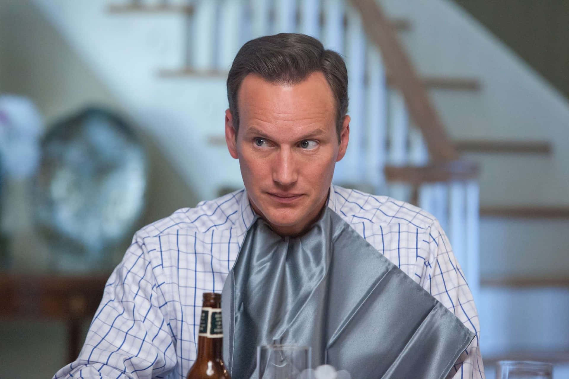 Patrick Wilson 桌布 壁紙 — 一個穿著格子襯衫的男人坐在桌旁，肩上披著一條銀色餐巾，旁邊放著一個啤酒瓶，表情嚴肅地望向鏡頭外。