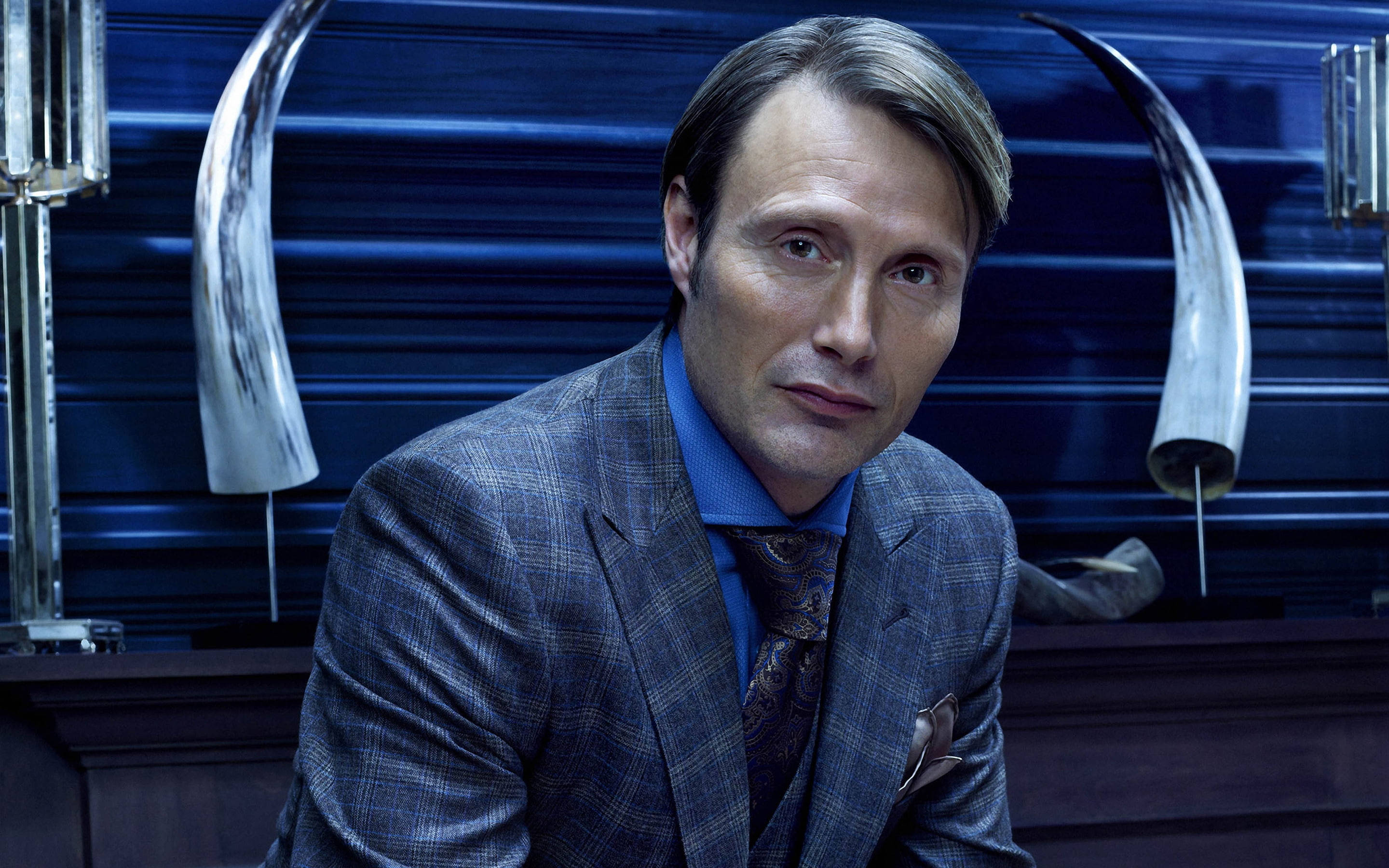 Mads Mikkelsen 桌布 壁紙 — 一個穿著格子西裝的男人自信地坐在藍色條紋牆前，牆上掛著角狀裝飾和獎盃。