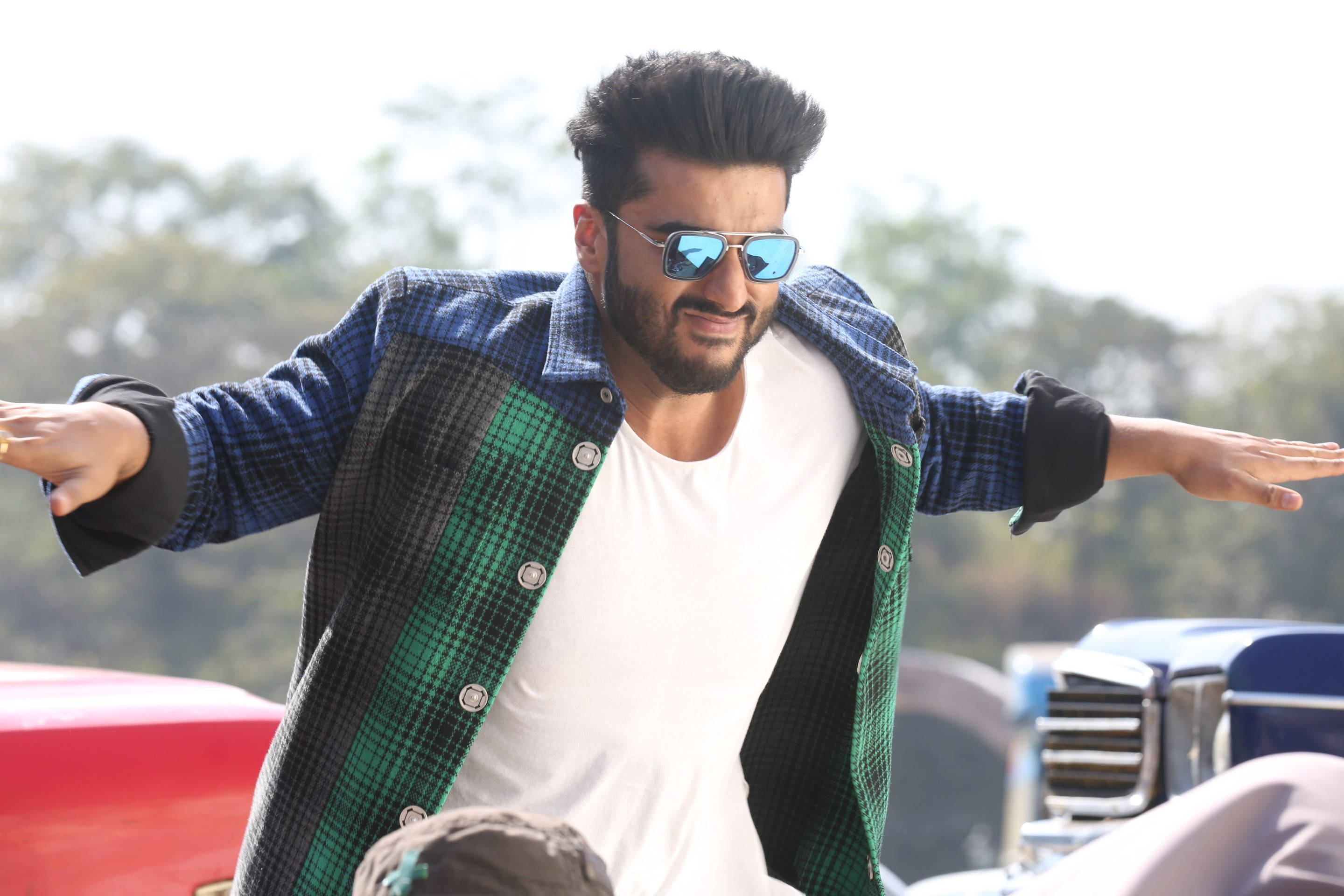 Arjun Kapoor 桌布 壁紙 — 一個穿著格紋外套和太陽眼鏡的男人，在戶外張開雙臂擺出自信的姿勢。