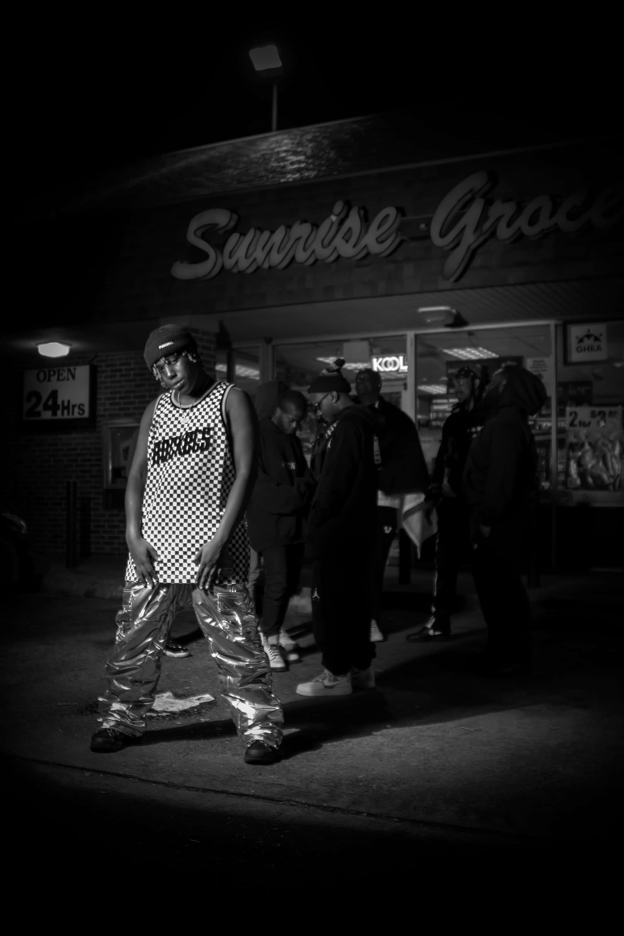 Don Toliver 桌布 壁紙 — 一個穿著格紋背心和閃亮褲子的男人在夜間自信地站在24小時營業的雜貨店外，在刺眼的人造燈光下。
