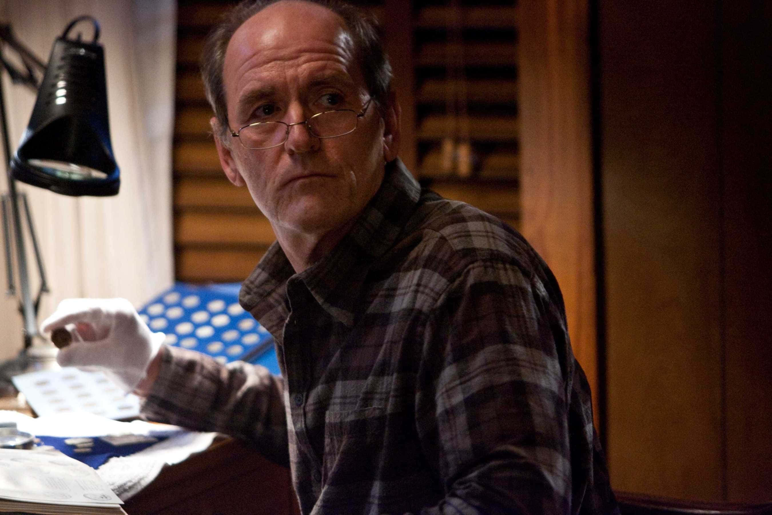 Richard Jenkins 桌布 壁紙 — 一個穿著格紋襯衫、戴眼鏡的男人，坐在黑色燈下雜亂的書桌前，表情嚴肅。