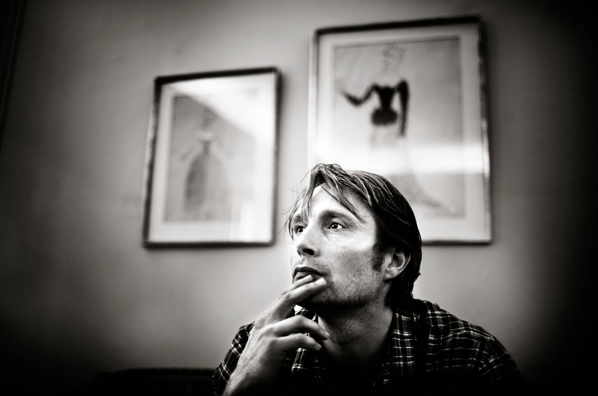 Mads Mikkelsen 桌布 壁紙 — 一個穿著格紋襯衫、沉思中的男人，手托下巴向上凝視，背景是柔和模糊的牆壁和帶框的藝術品。