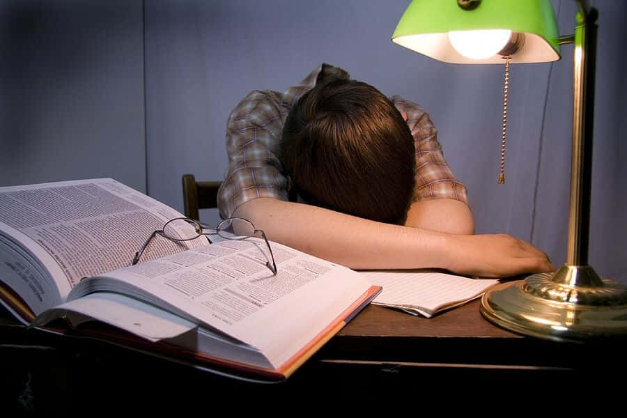 Studying 桌布 壁紙 — 一個穿著格紋襯衫的人在桌上睡覺，旁邊有一本打開的書、眼鏡和一盞發出溫暖光線的綠色燈。