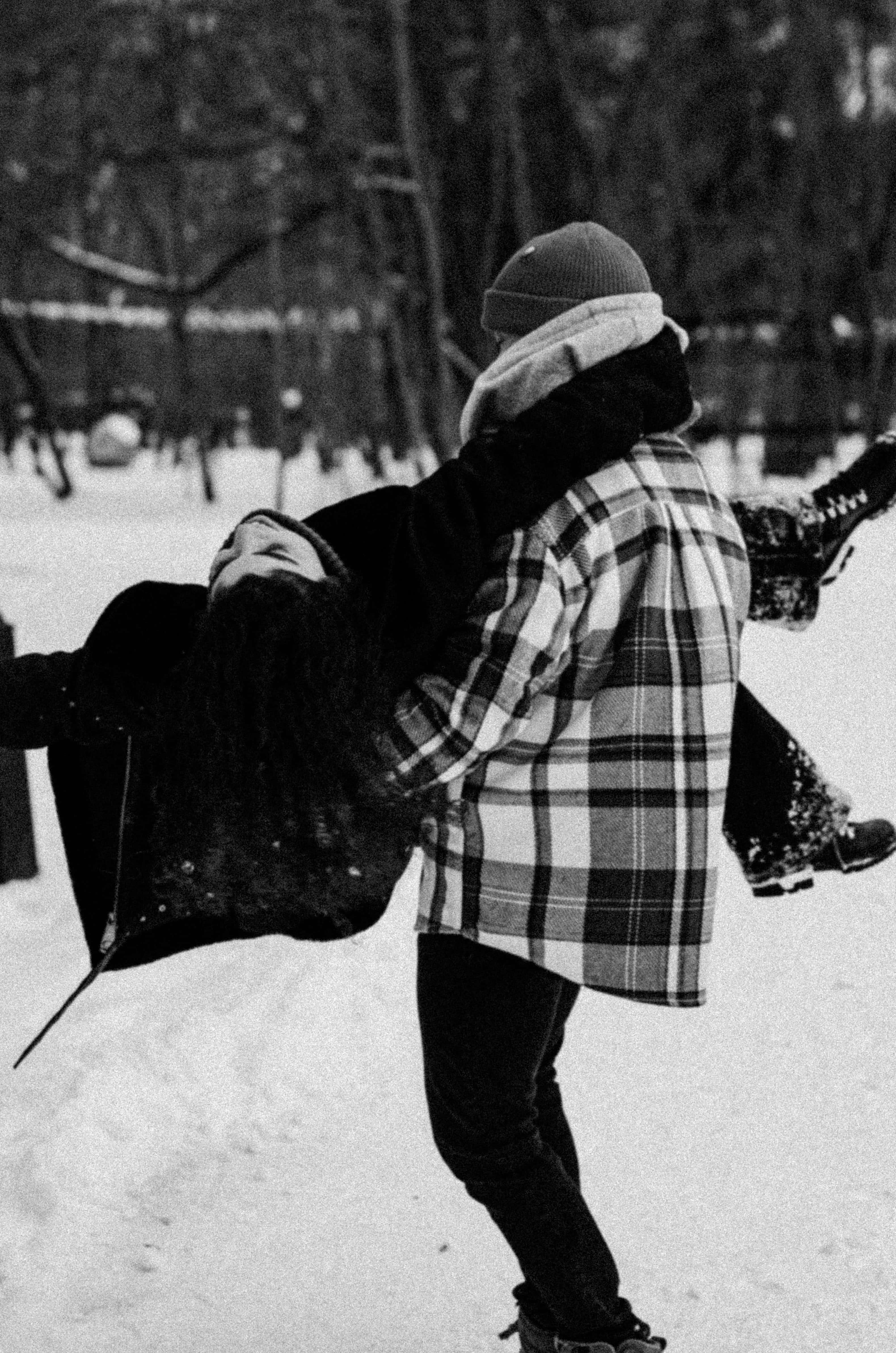 黑白 桌布 壁紙 — 一個穿著格紋襯衫的人在白雪皚皚的冬季公園中，在光禿禿的樹木之間，以黑白畫面開心地把另一個人舉起。