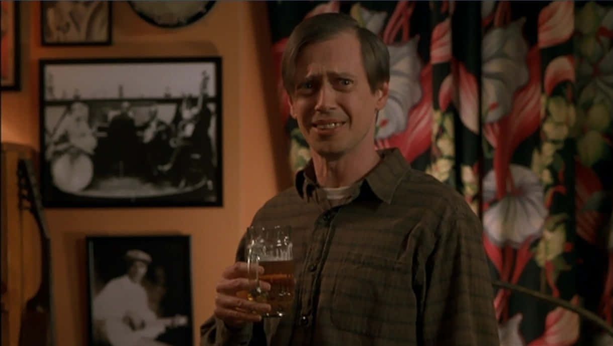 steve buscemi 桌布 壁紙 — 一個穿著格紋襯衫的男人在燈光溫暖的房間裡，手裡拿著啤酒杯，房間有花卉圖案窗簾和牆上的相框。