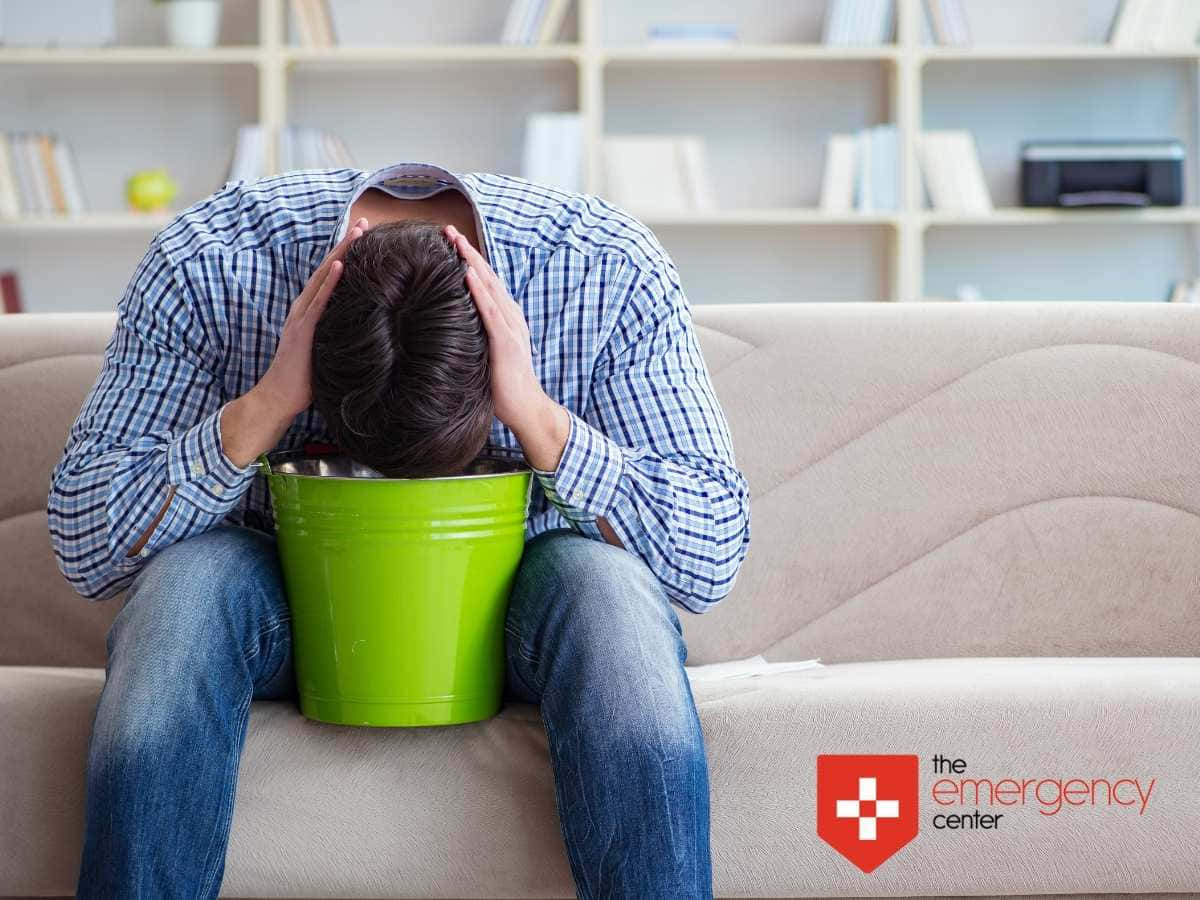 生病 桌布 壁紙 — 一個穿著格紋襯衫的男人坐在米色沙發上，頭埋在一個亮綠色水桶裡，雙手放在頭上，房間裡有書架。