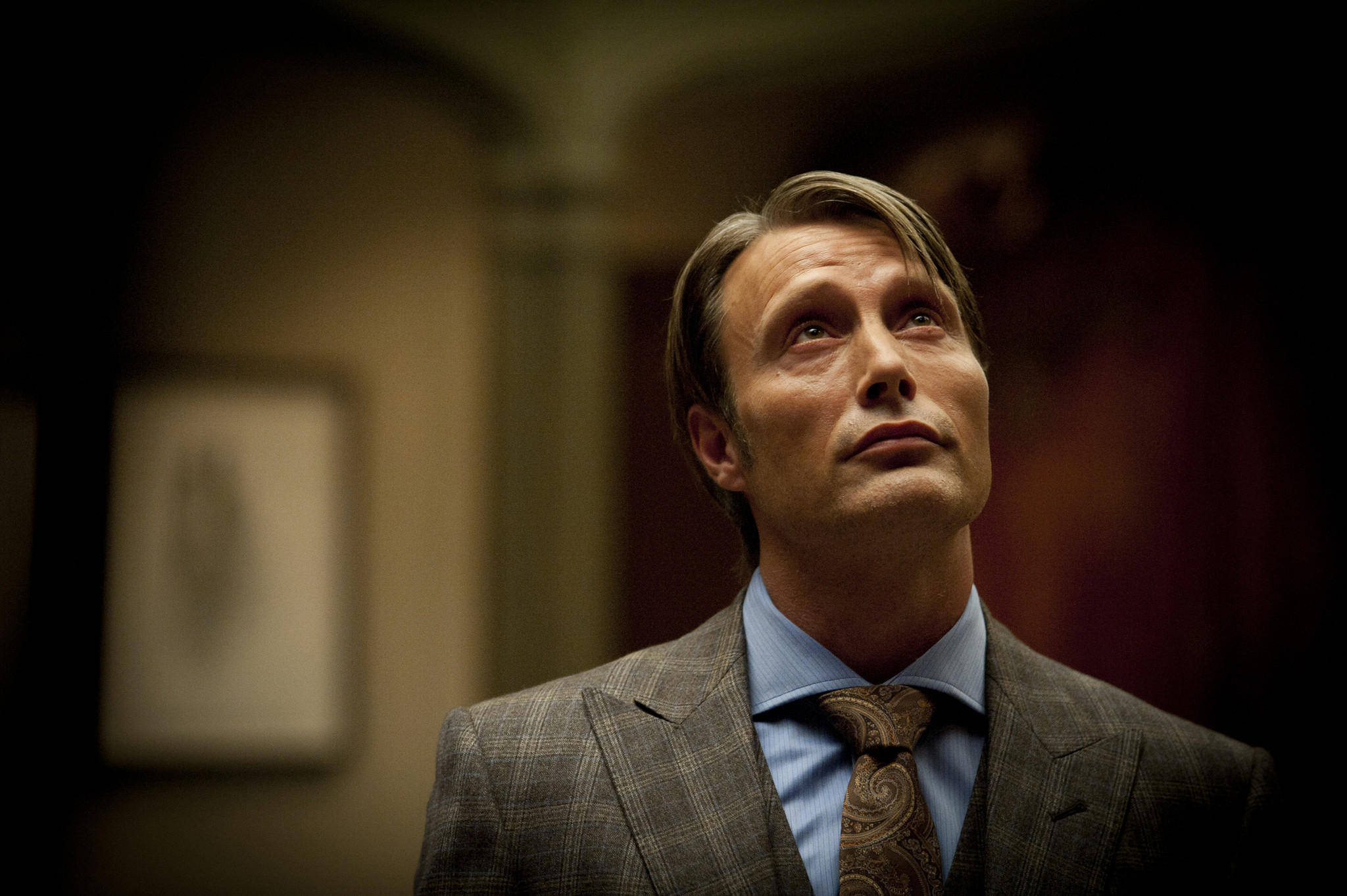 Mads Mikkelsen 桌布 壁紙 — 一個穿著格紋西裝和佩斯利領帶的男人在昏暗的房間裡嚴肅地向上看。
