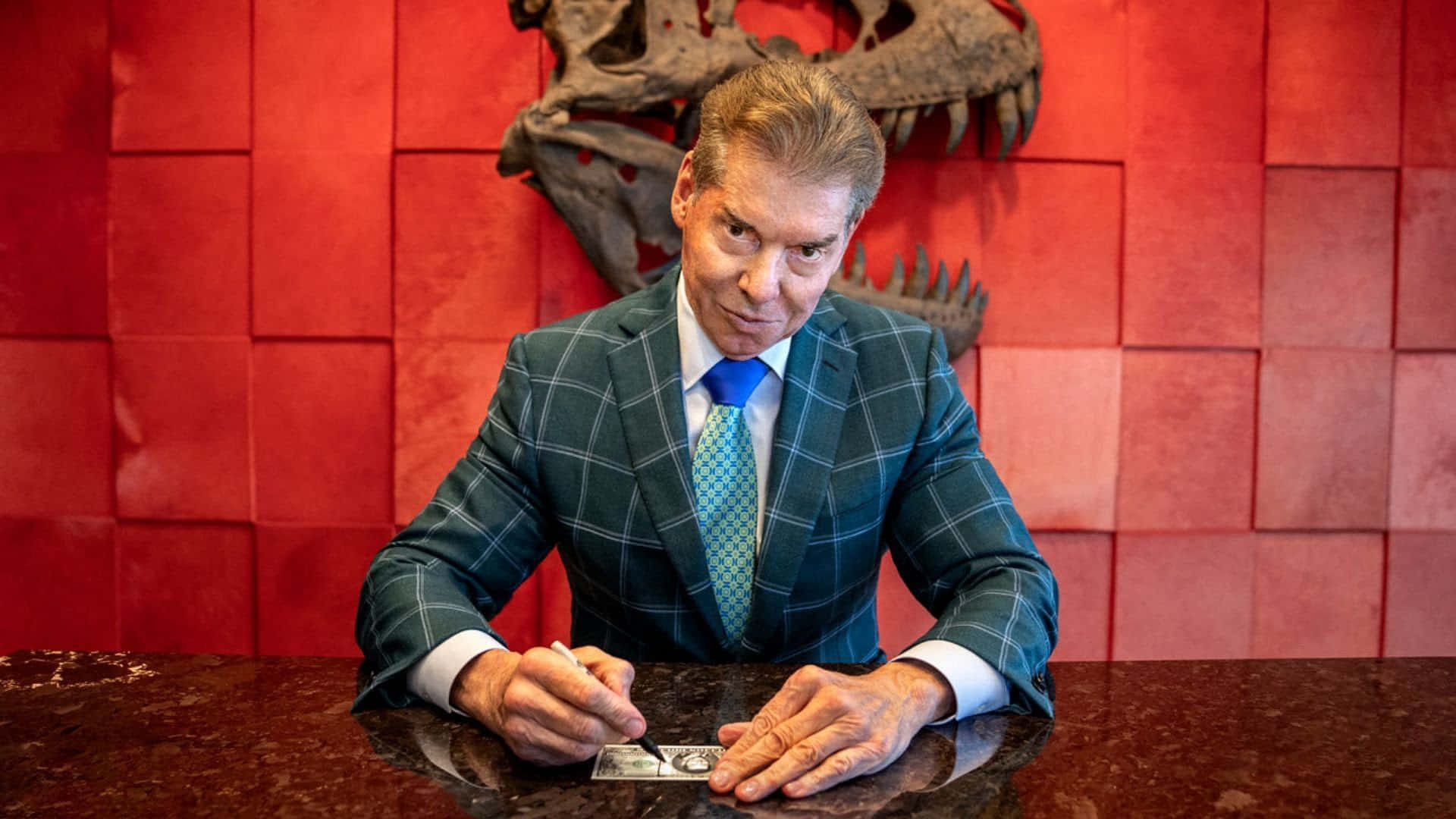WWE 桌布 壁紙 — 一個穿著格紋西裝的男人在一張深色大理石桌上簽署一張一美元紙幣，背景是紅色瓷磚牆，上方有一個恐龍頭骨。