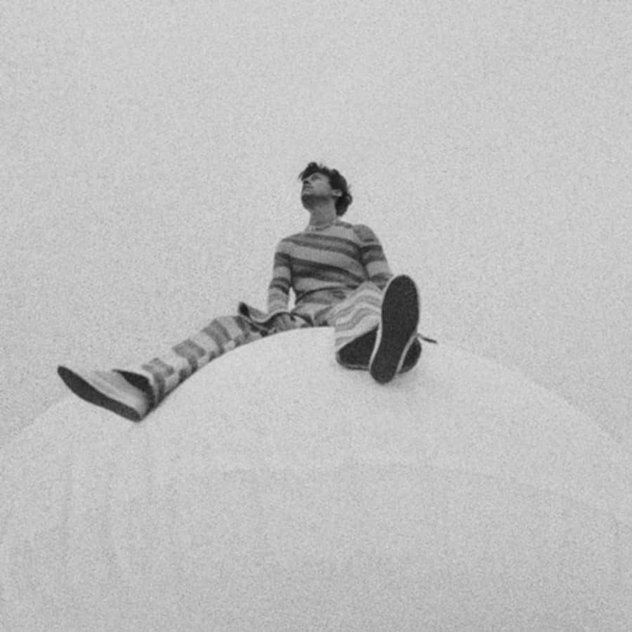 Harry Styles Black And White 桌布 壁紙 — 一個穿著條紋衣服的人坐在彎曲的白色表面上，仰望朦朧的單色天空。