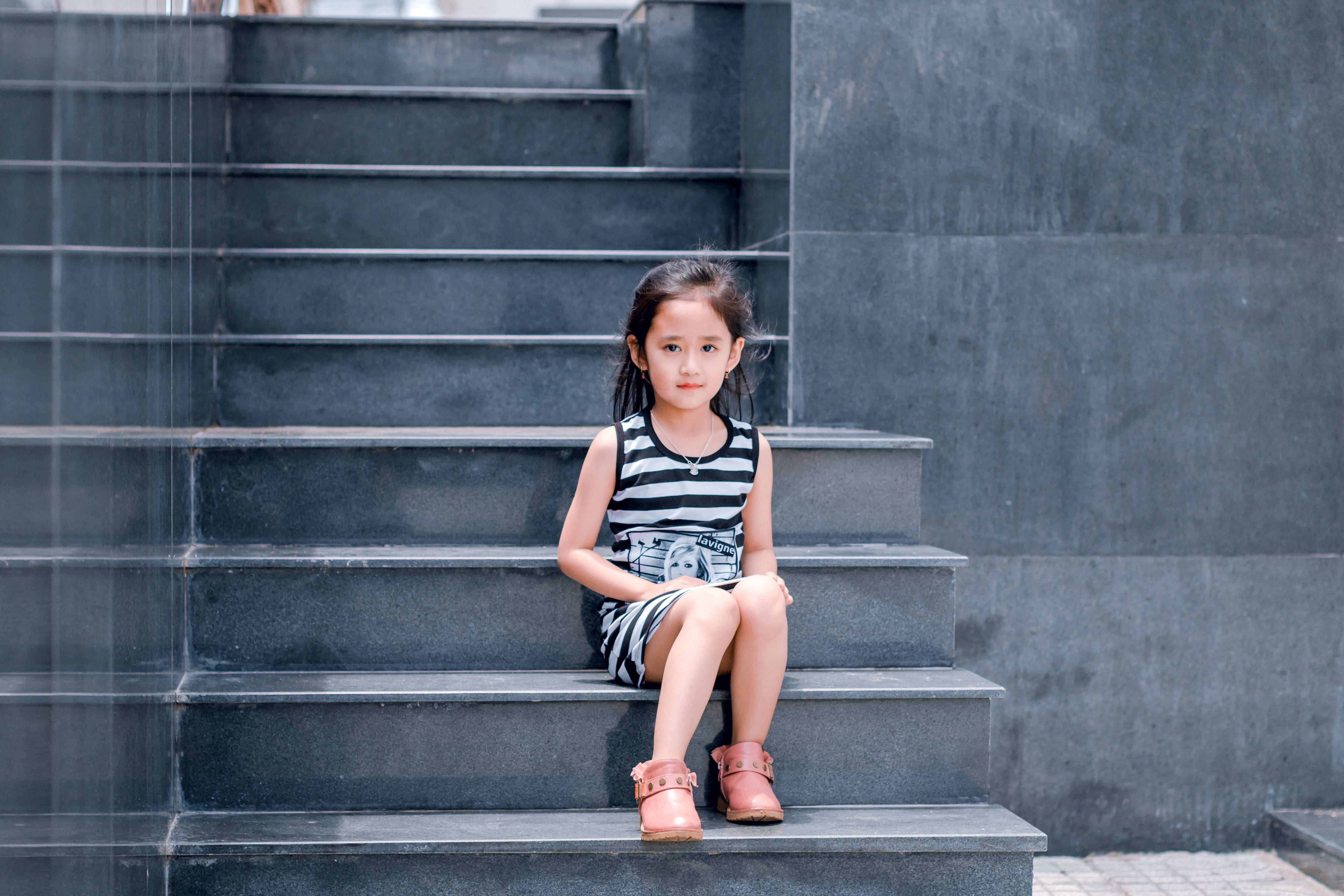 小孩動圖 桌布 壁紙 — 一個穿著條紋裙的小女孩平靜地坐在深灰色混凝土樓梯上，背景是現代風格的牆壁。