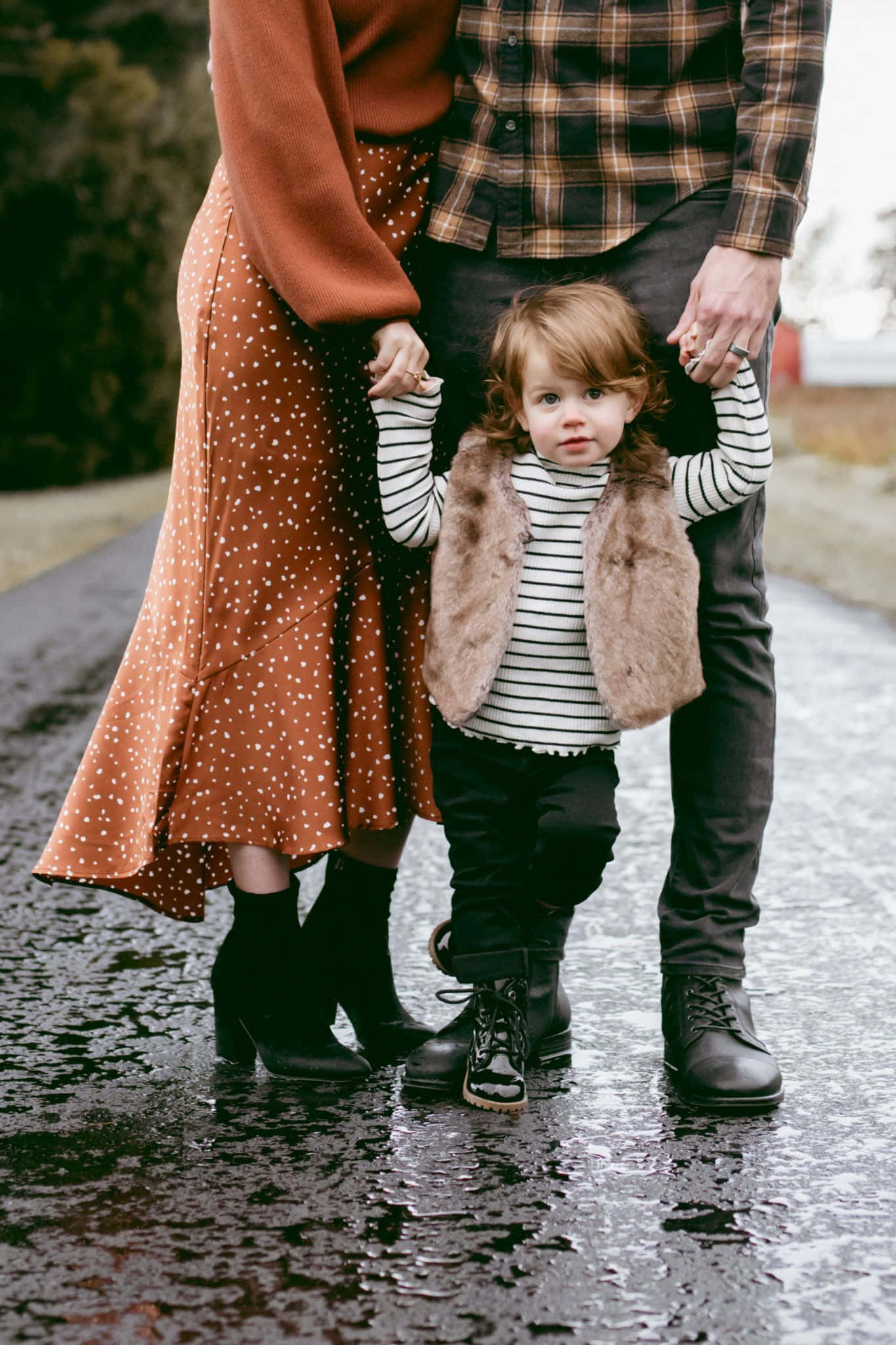 Family 桌布 壁紙 — 一個穿著條紋襯衫和仿皮草背心的幼兒站在濕漉漉的人行道上牽手的父母之間，直視鏡頭。