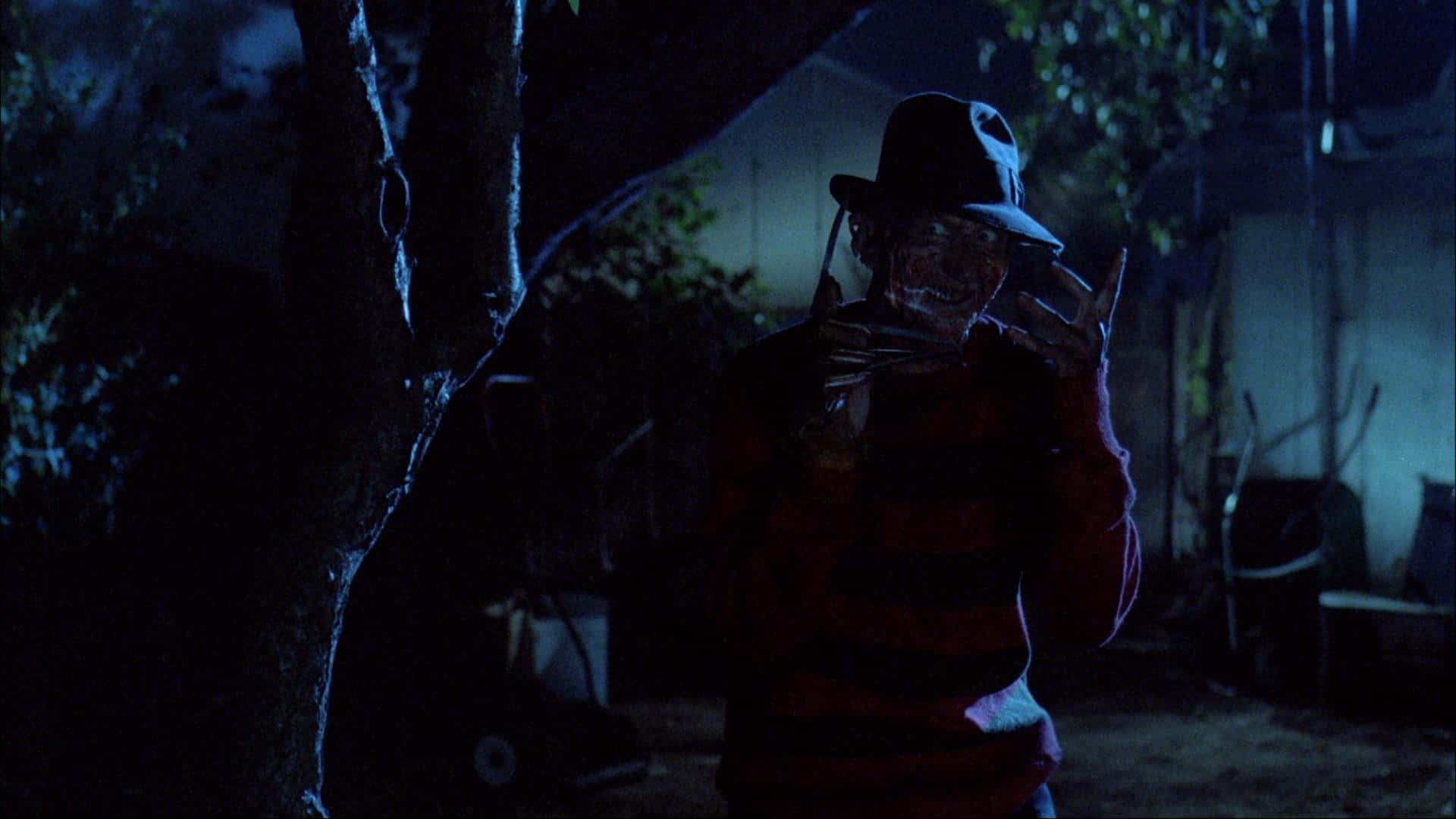 Freddy Krueger 桌布 壁紙 — 一個穿著條紋襯衫和帽子的可怕人物，夜晚站在黑暗的後院，手持刀，眼睛發光。