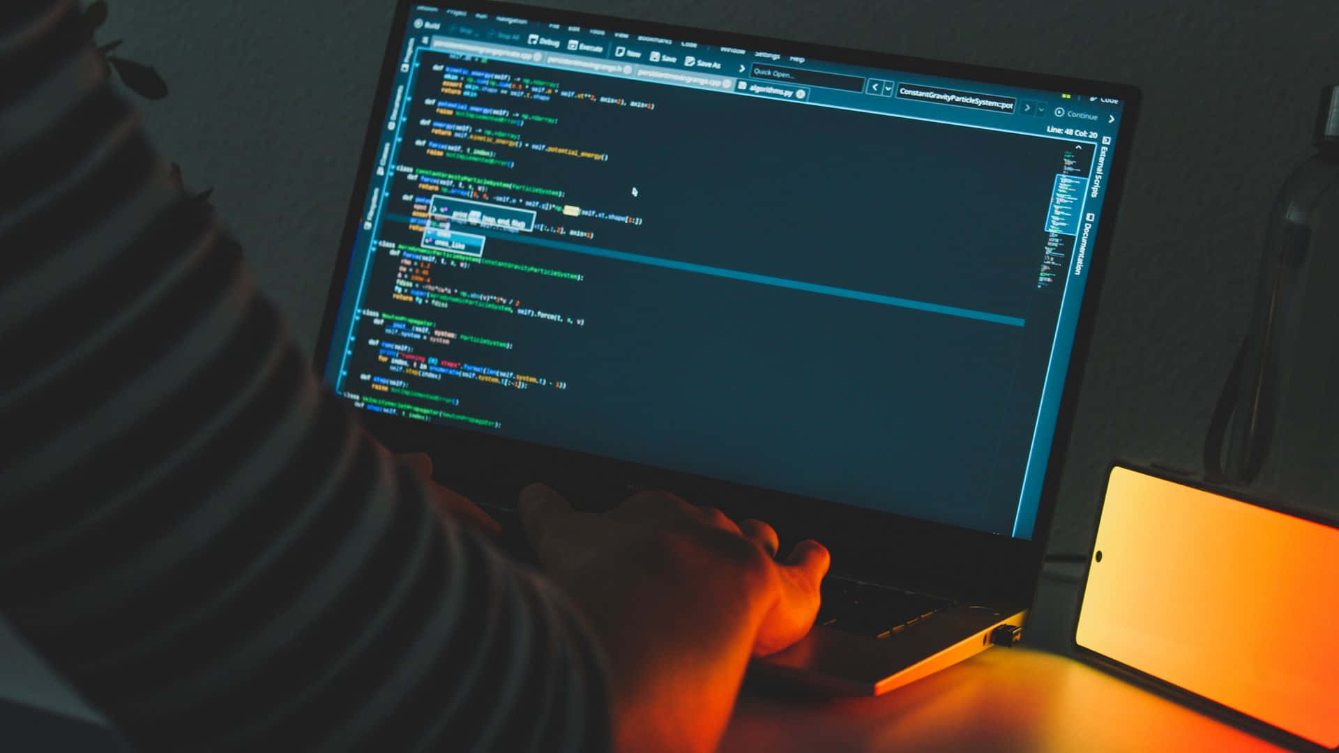 Code 桌布 壁紙 — 一個穿著條紋襯衫的人在溫暖的環境燈光下，在發光螢幕的筆記型電腦上打字寫程式碼。