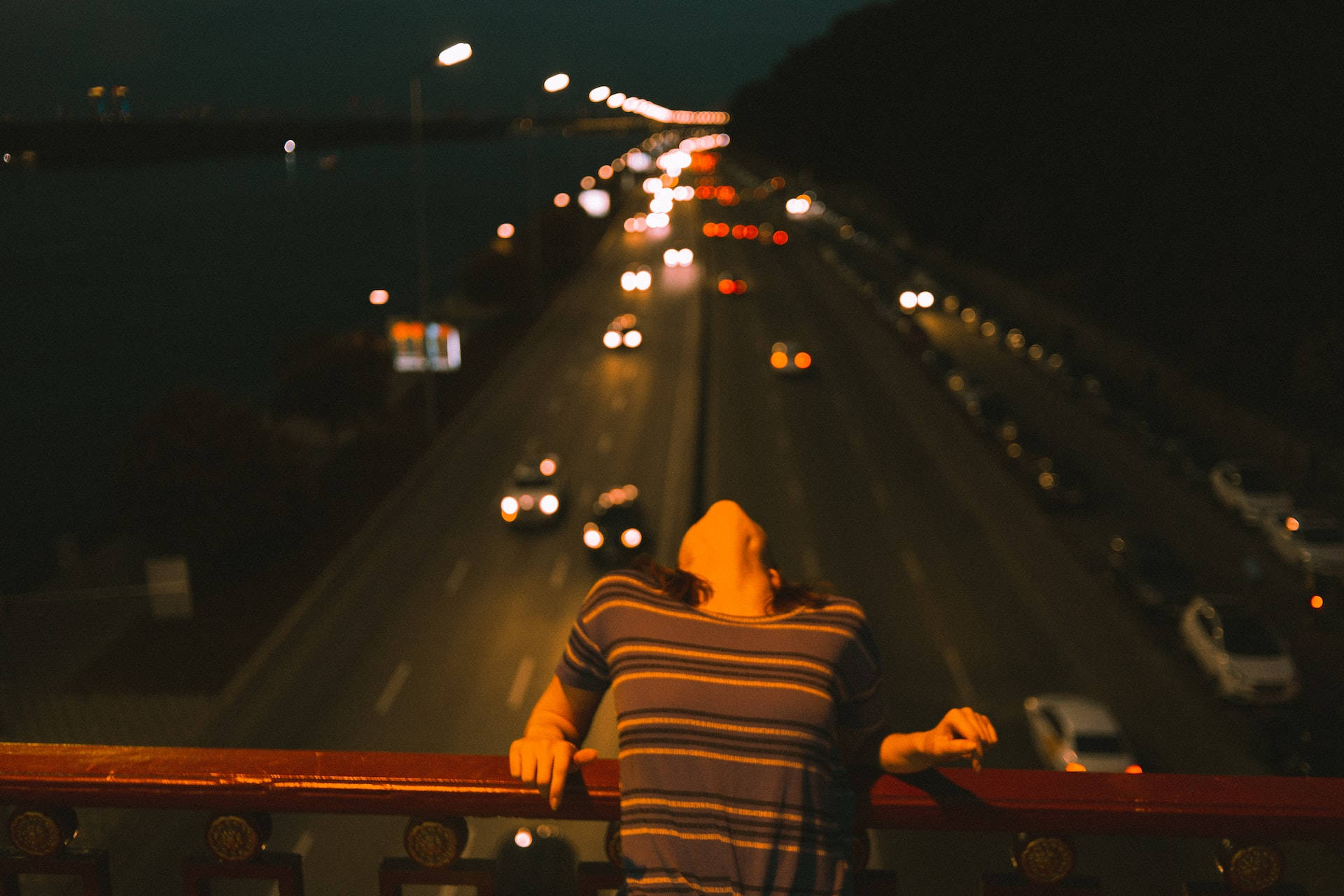 Night Aesthetic 桌布 壁紙 — 一個穿著條紋襯衫的人夜晚靠在欄杆上，俯瞰著一條車水馬龍、車燈閃爍的高速公路。